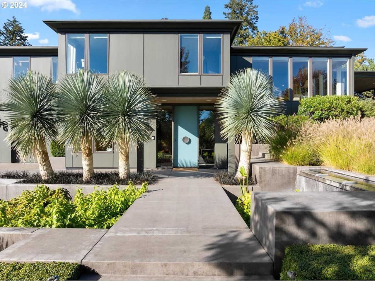 Modern gray house with turquoise door and palm trees. Concrete walkway and landscaping.