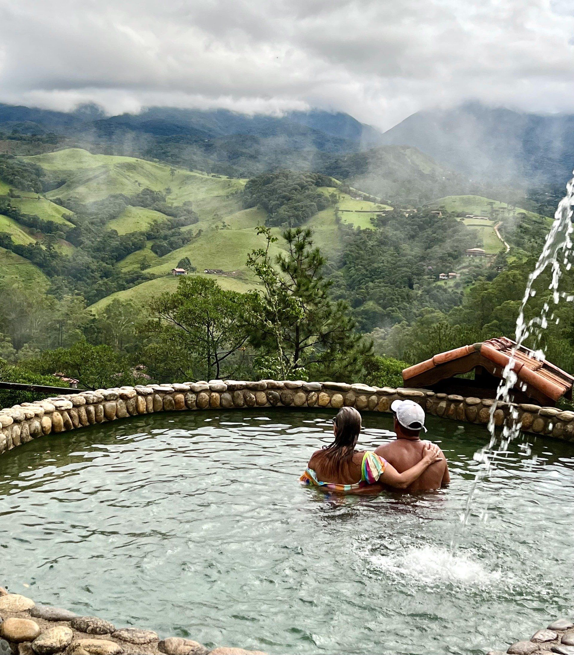 Um homem e uma mulher estão sentados em uma piscina com montanhas ao fundo