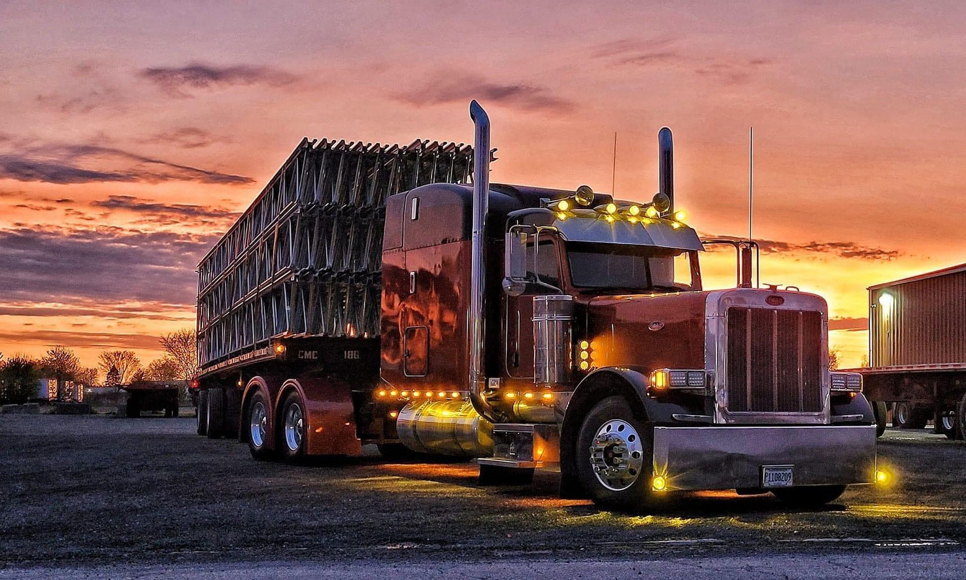 Red semi-truck with a large load of goods, parked against a colorful sunset sky.