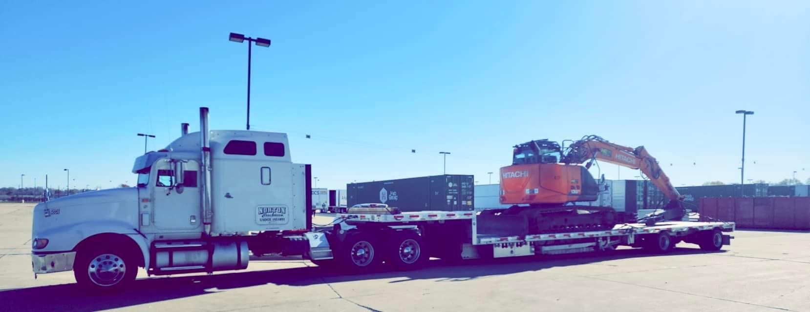 A white semi-truck hauling a flatbed trailer with an excavator and shipping containers on a sunny day.
