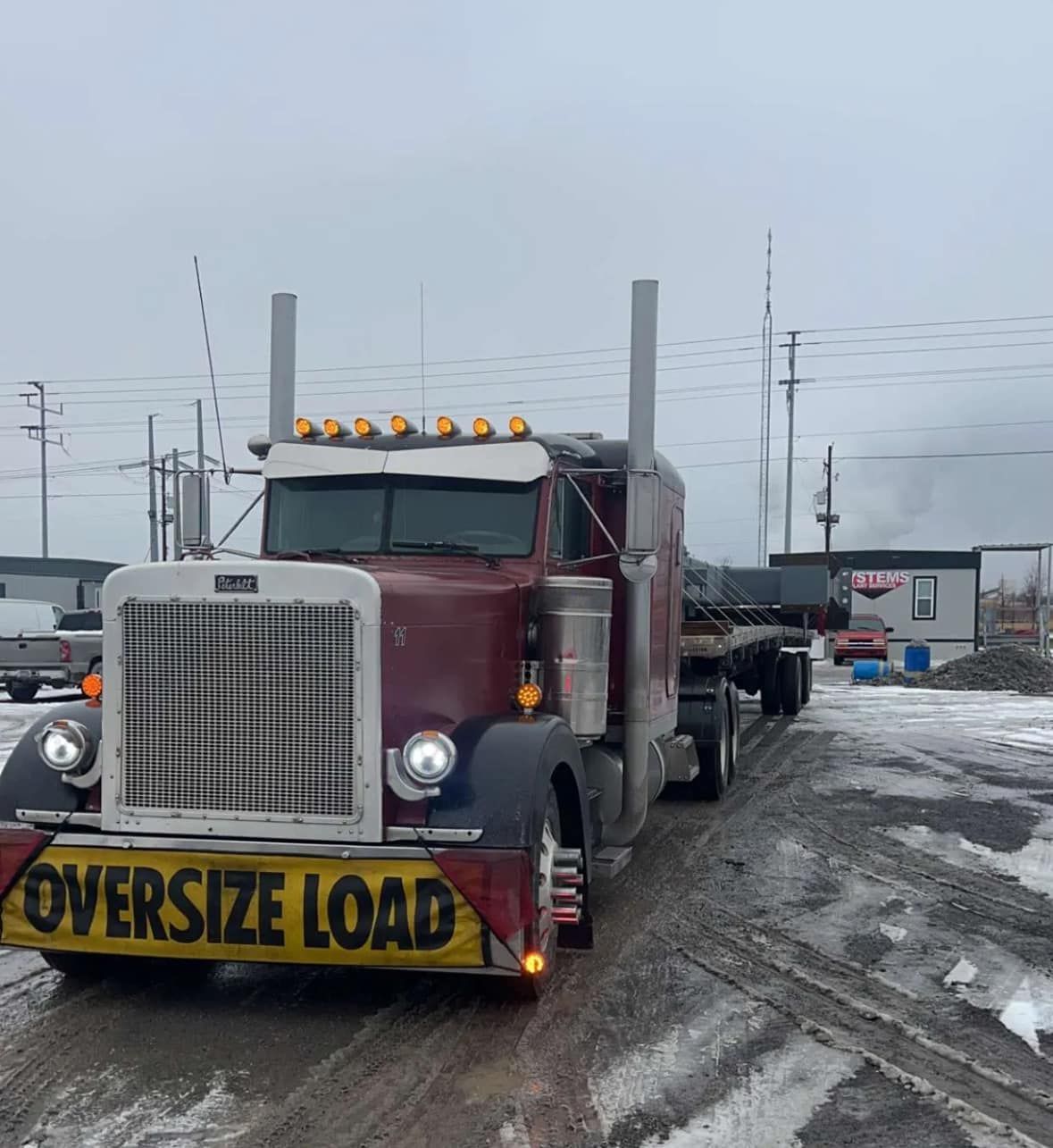 Burgundy Peterbilt truck hauling an oversized load on a flatbed trailer; snowy, industrial setting.