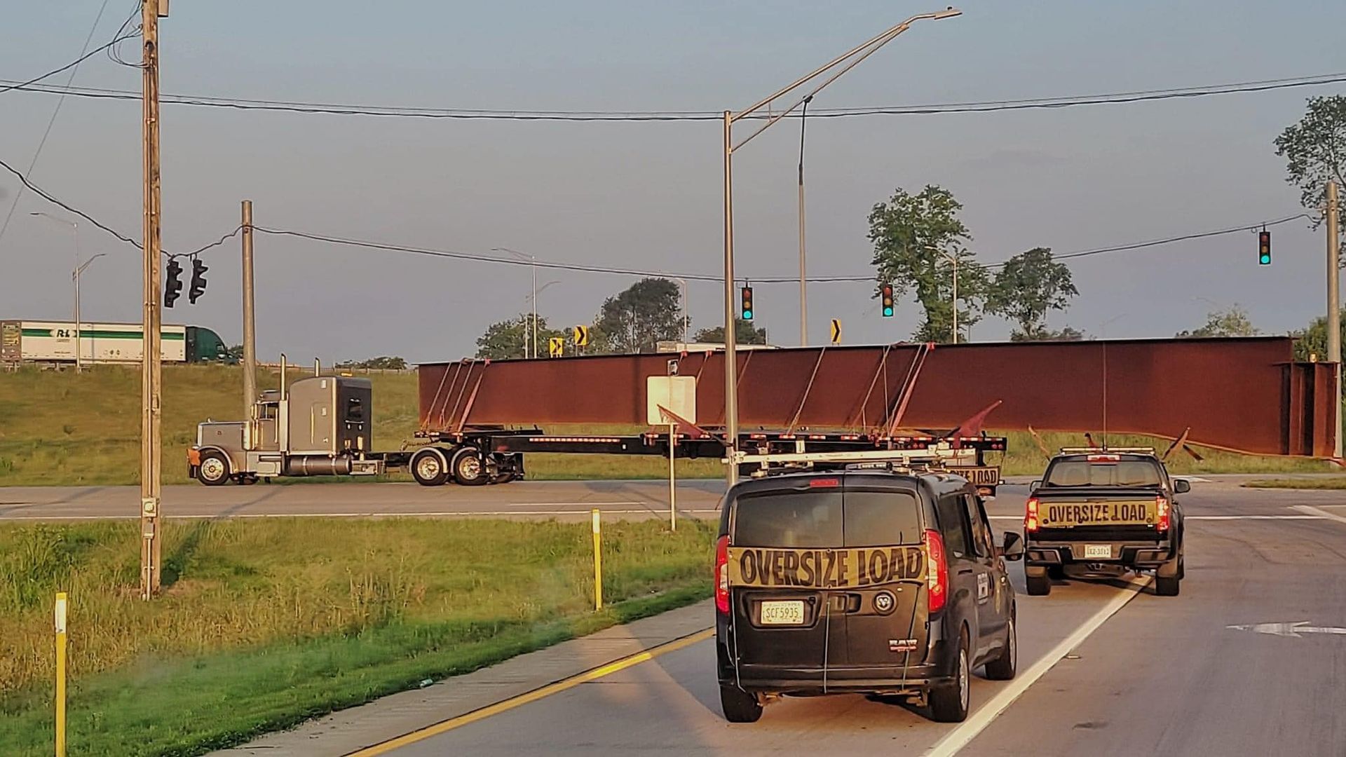 A flatbed truck carrying a long, brown steel beam on a road, with vehicles and power lines visible.