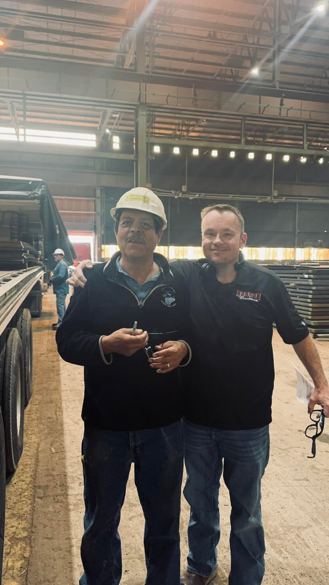 Two men in a warehouse: one in a hard hat, the other with an arm around him. They are smiling near a truck trailer.