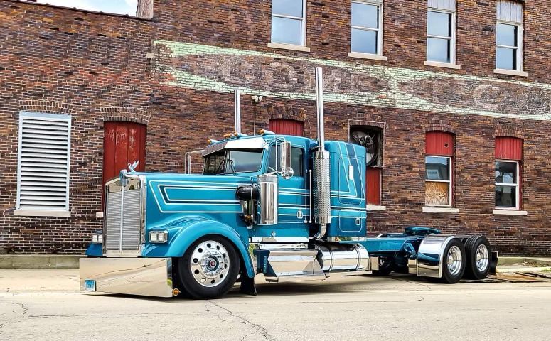 Turquoise semi-truck parked in front of a brick building with silver accents and chrome exhaust pipes.