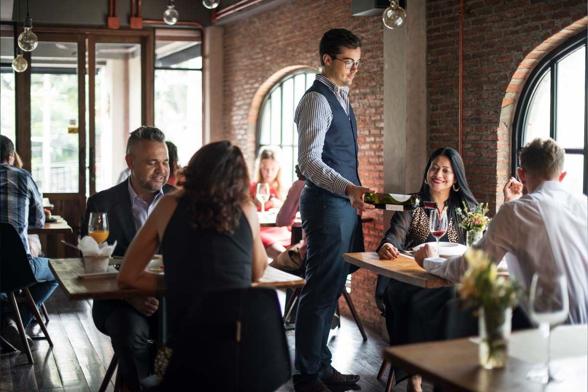 Waiter pouring wine at a restaurant table with several seated guests. Brick walls and arched windows.