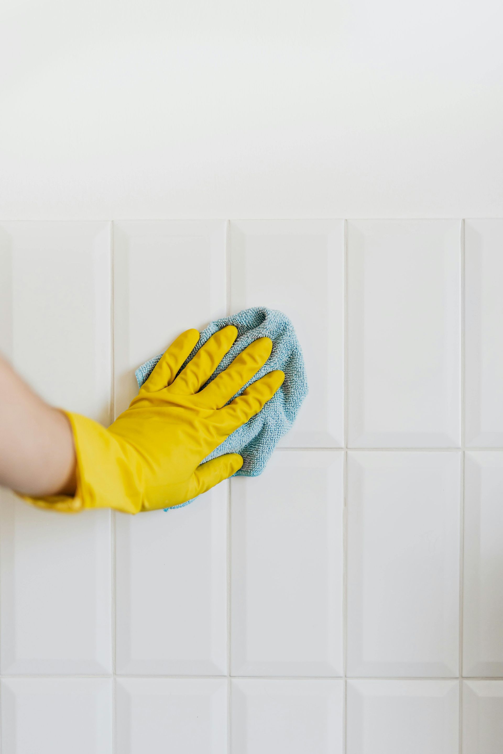 A person wearing yellow gloves is cleaning a white tile wall with a sponge.