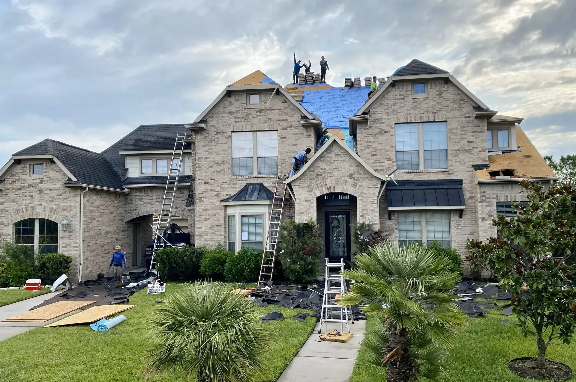 Workers perform roofing repairs on a large brick two-story house with blue protective underlayment on the roof.