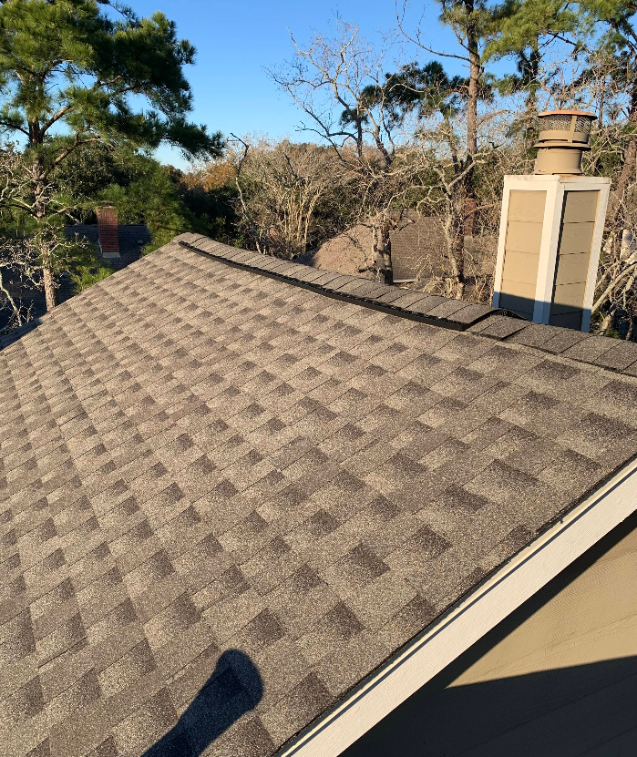 A high-angle view of a residential shingled roof with a chimney, framed by trees against a clear blue sky.
