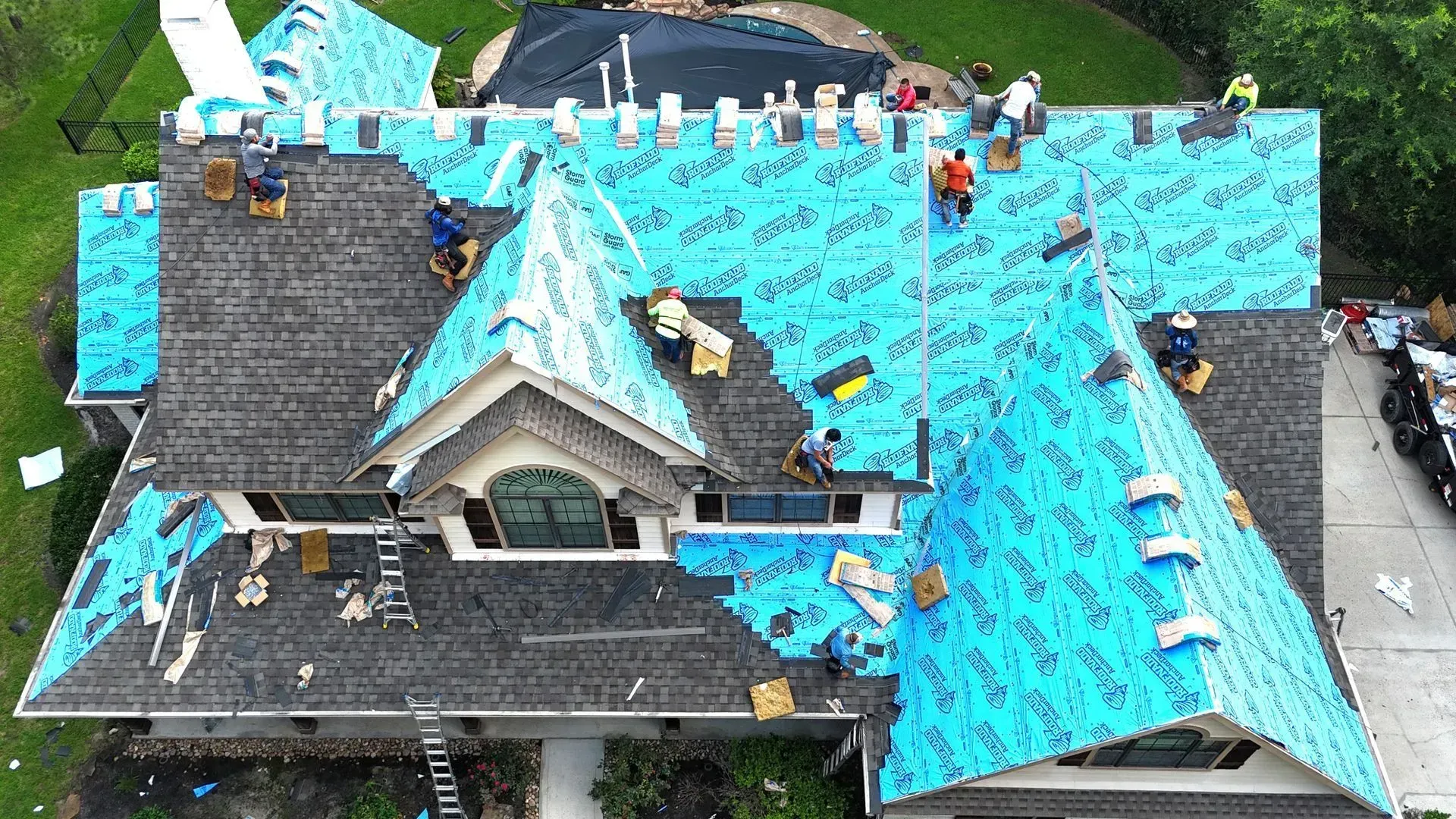 Aerial view of a team of roofers installing new roofing shingles on a house partially covered with blue underlayment.