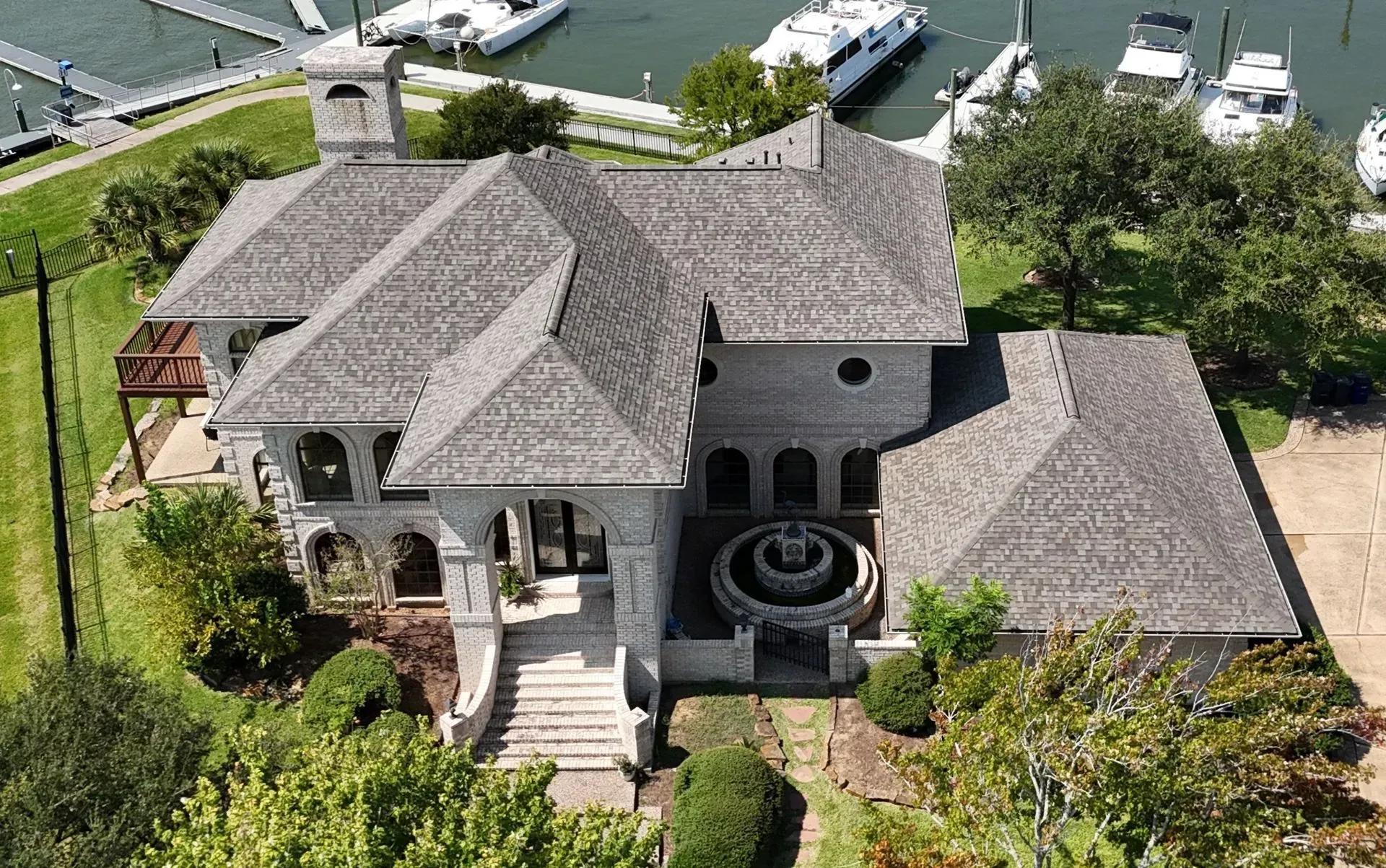 Aerial view of a large, light-colored waterfront house with a grey shingled roof, a fountain, and nearby boat docks.