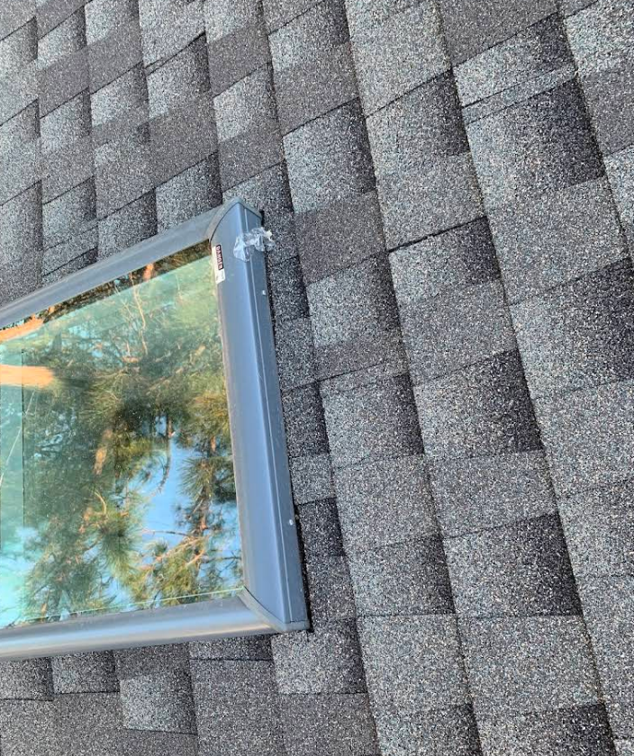 A gray-framed skylight installed on a dark shingled roof, reflecting blue sky and tree branches.