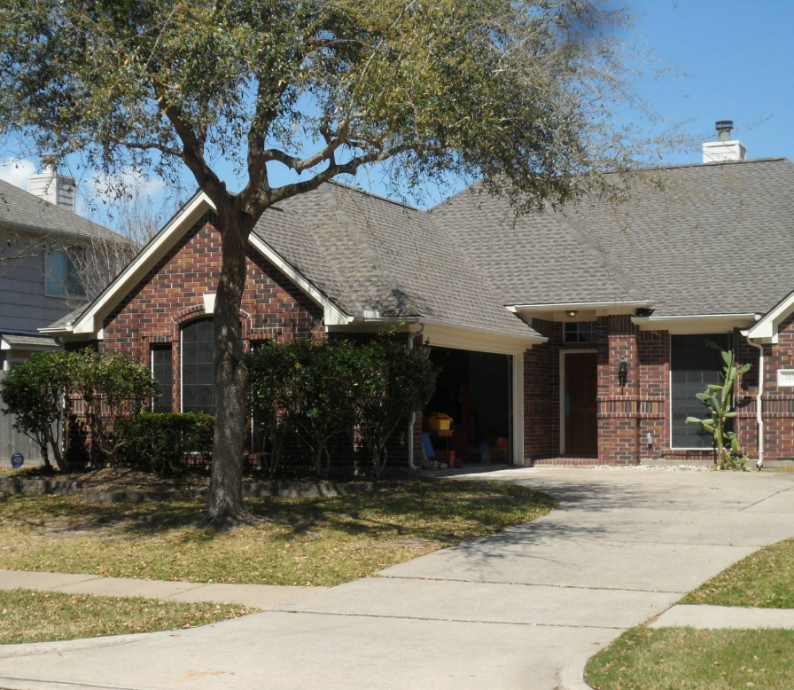 A one-story brick house with a dark shingled roof, a paved driveway, and a large tree in the front yard.