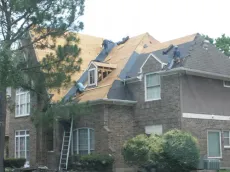 Workers install shingles on the roof of a two-story brick house with a ladder leaning against the front.