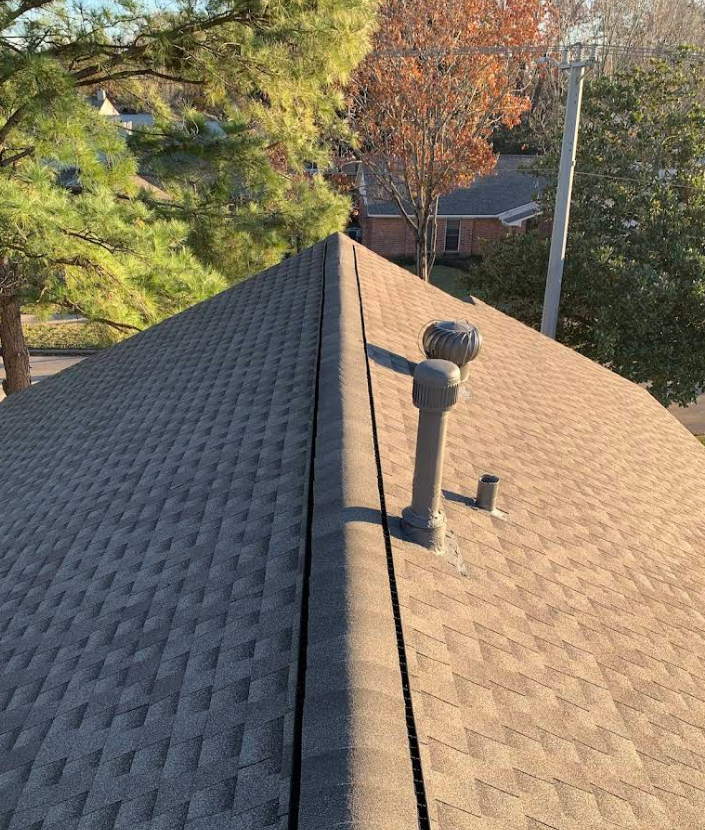 A view of a roof ridge featuring brown shingles, two vent pipes, and surrounding trees in a residential neighborhood.