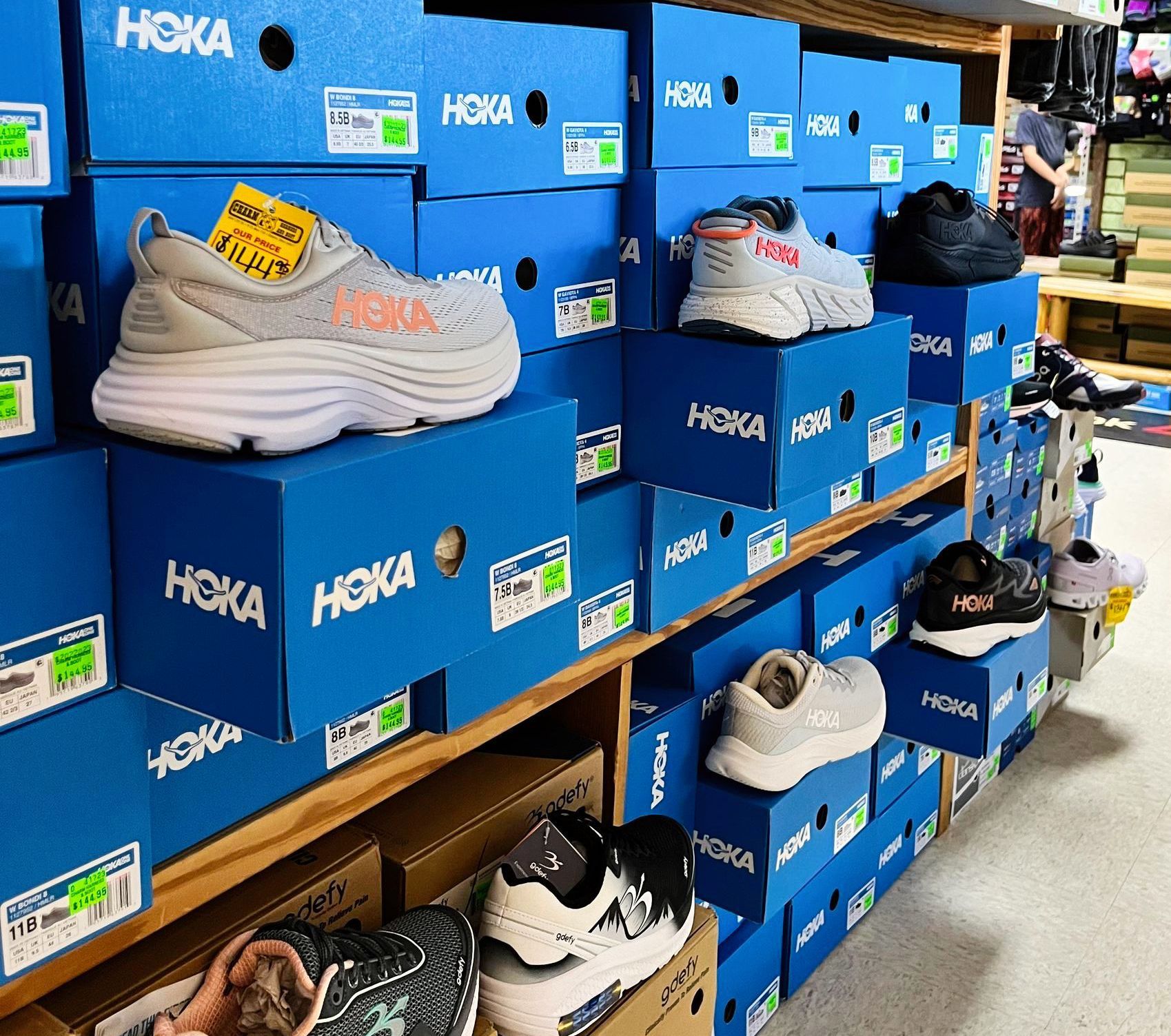 Shoes on blue HOKA boxes in a store; various colors.