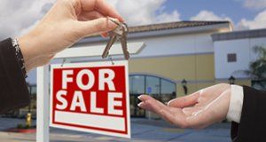 An outstretched hand receiving keys from another hand, with a red For Sale sign in the background