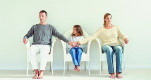 A couple sitting down, holding hands across a child who is sitting between them; all in white tub chairs