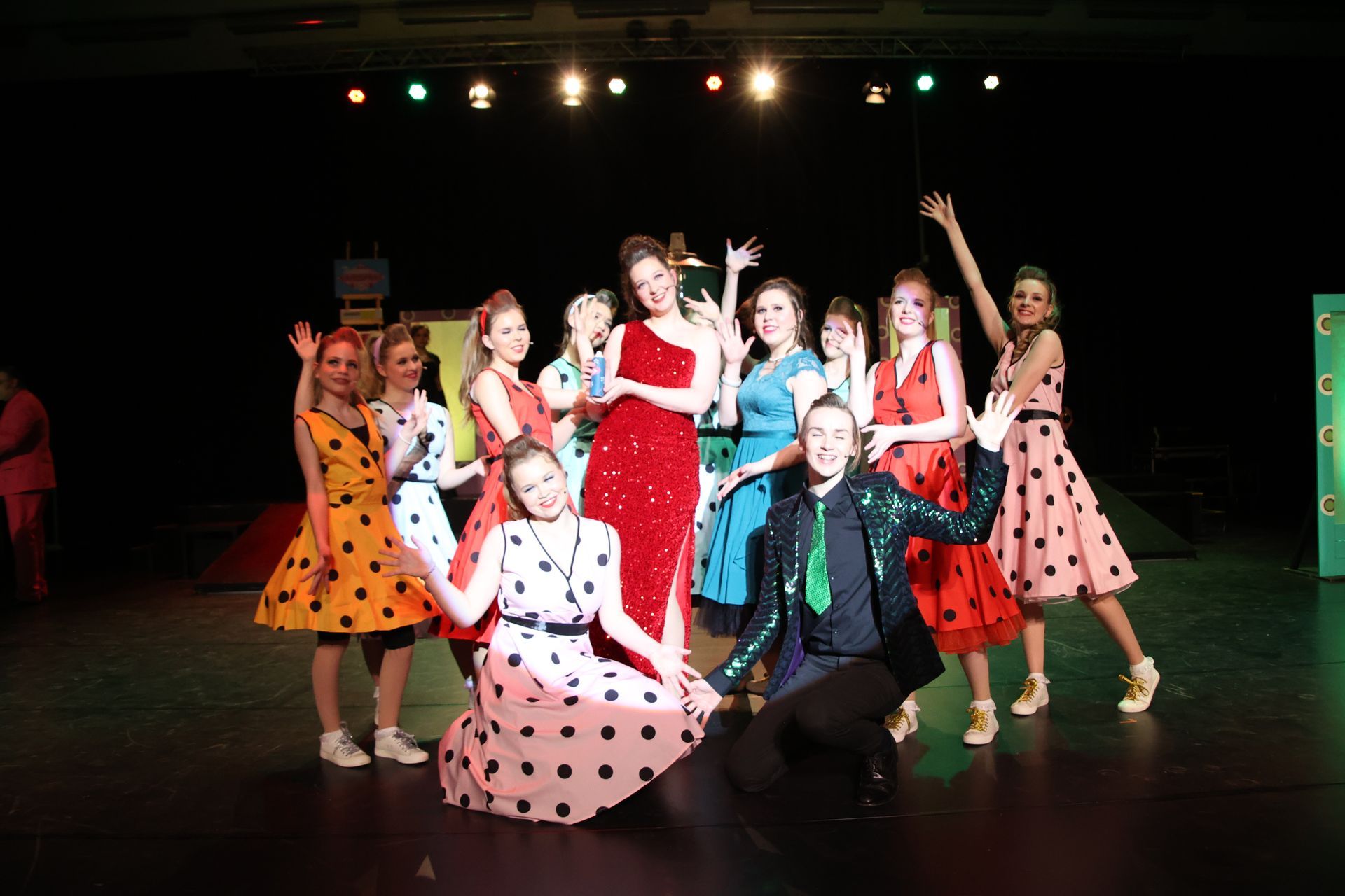 Een groep artiesten in kleurrijke jurken en pakken staat op het podium te zwaaien.