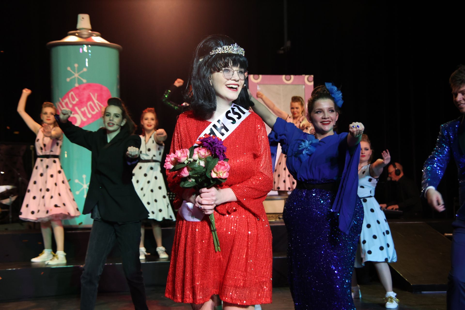 Een vrouw in een rode jurk en tiara staat op het podium met bloemen in haar hand, samen met danseressen in jurken met stippen.