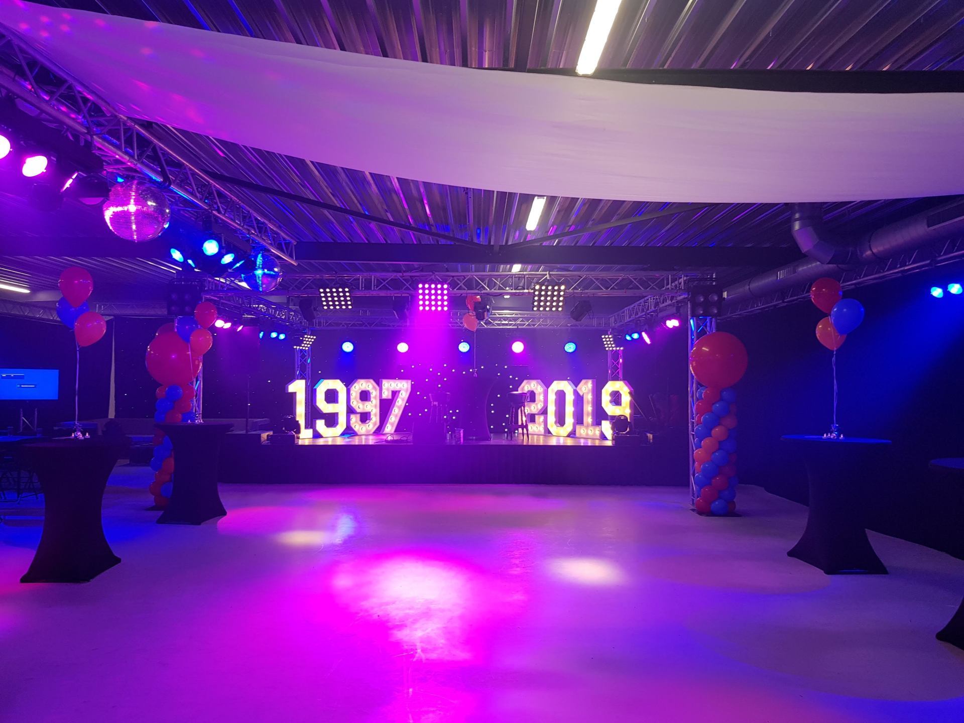 De evenementenruimte is versierd met paarse en blauwe verlichting, op het podium staat achter het achterdoek 