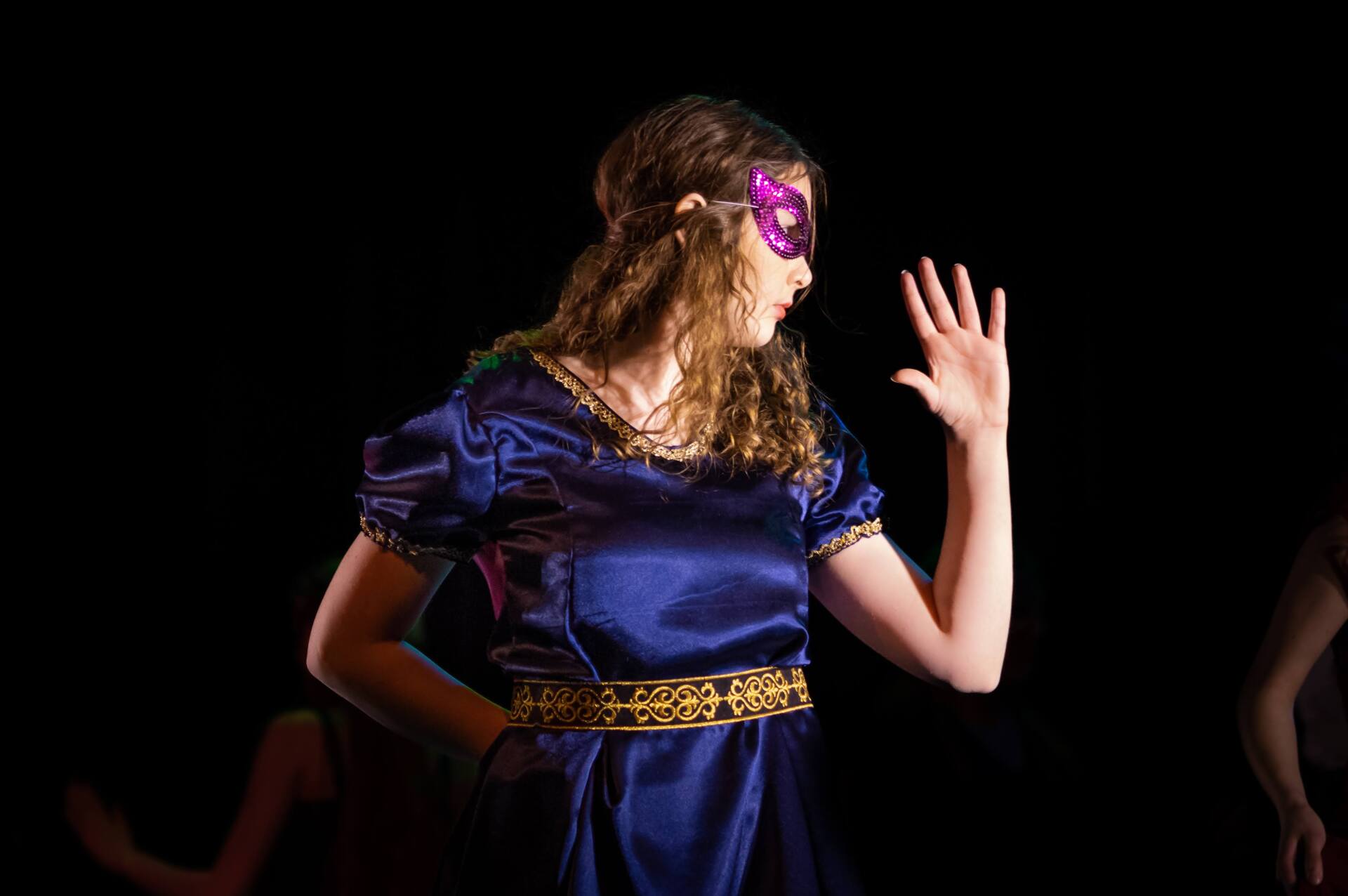 Vrouw in blauwe jurk en masker, gebarend met de hand, tegen een zwarte achtergrond.