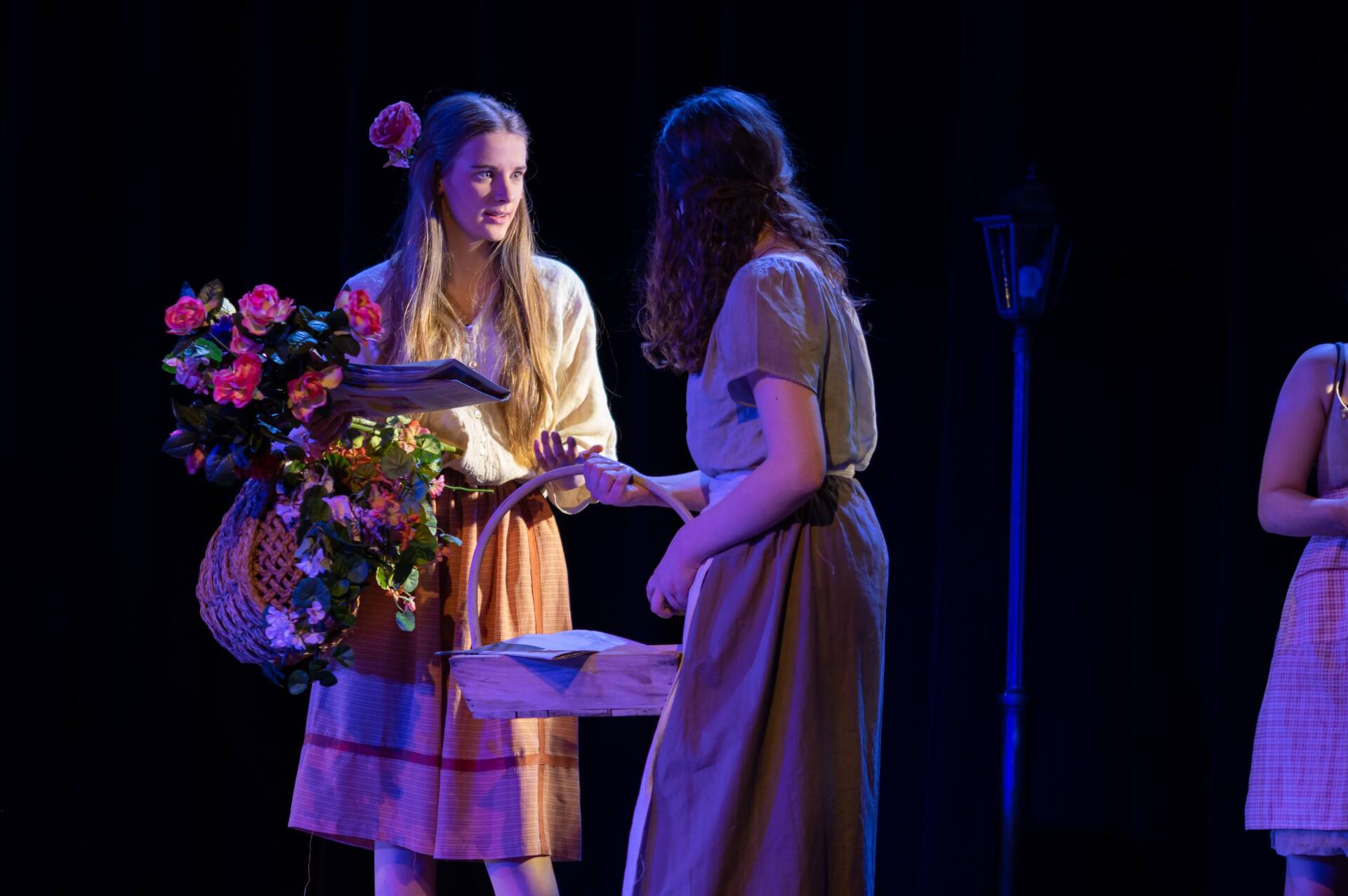 Twee jonge vrouwen op een podium, een van hen biedt bloemen aan, beiden in historische kostuums, donkere achtergrond.