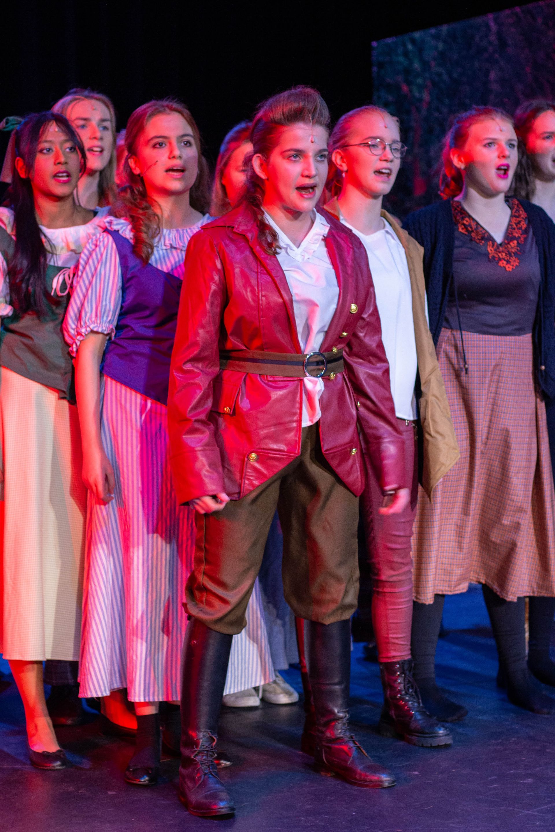 Een persoon in een rode jas en laarzen zingt op het podium met een koor van mensen in kleurrijke kostuums.