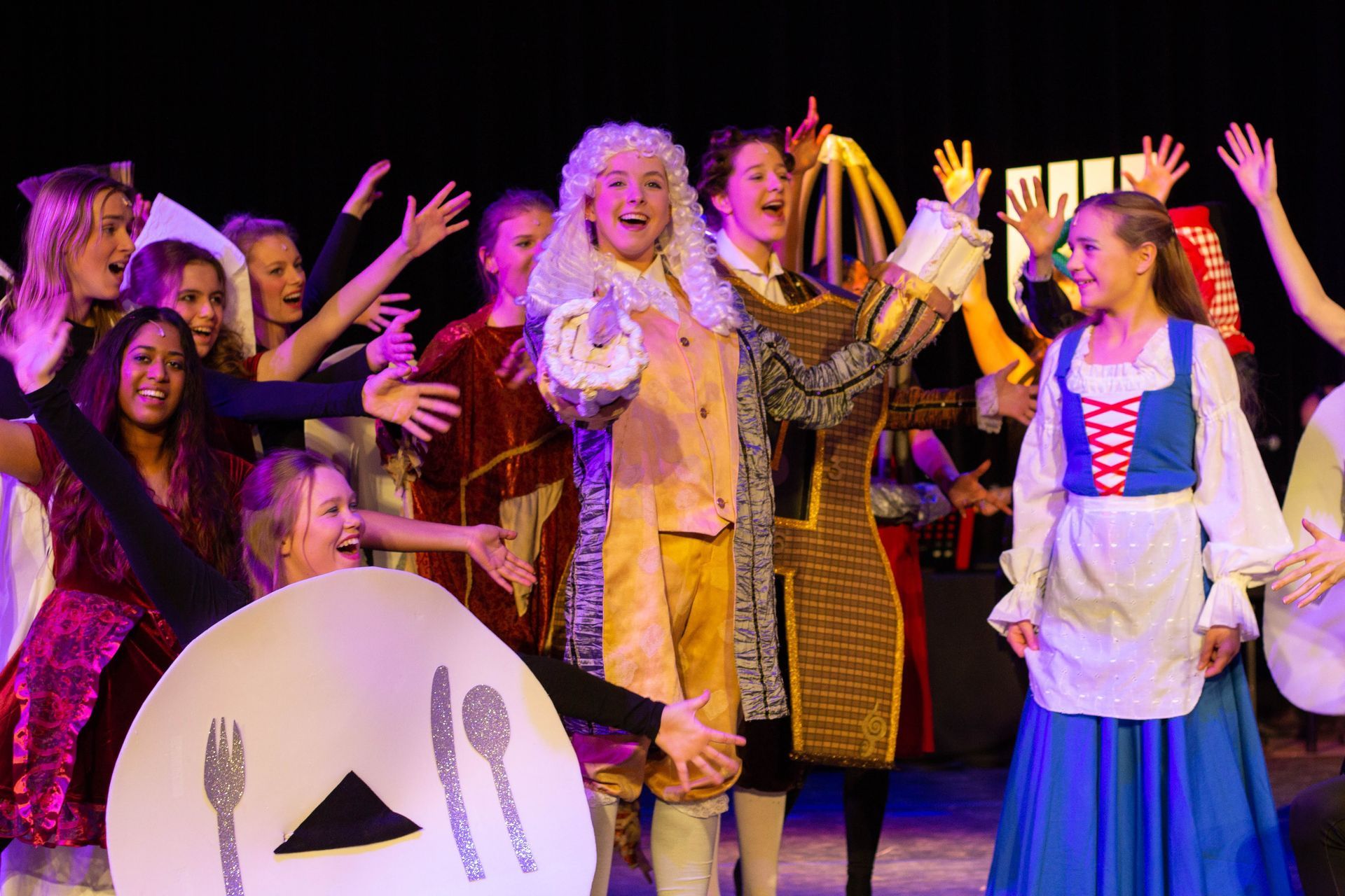 Een groep mensen in kostuums staat op het podium en speelt een toneelstuk. De hoofdrolspeler draagt ​​een witte pruik; de anderen hebben hun armen omhoog in een vrolijke setting.