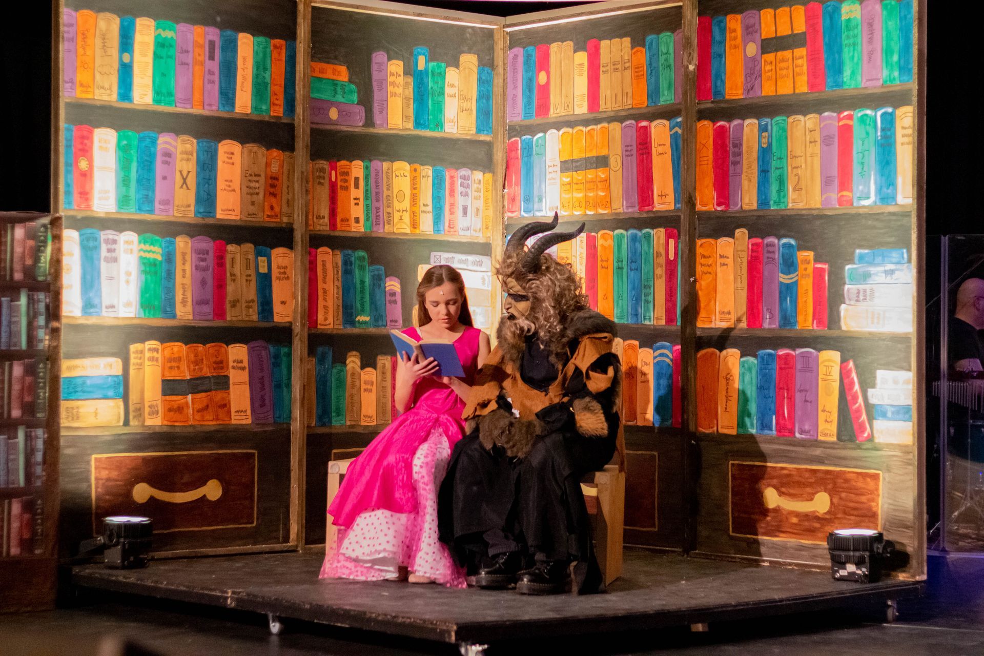 Belle en het Beest zitten te lezen voor een kleurrijke boekenplank als achtergrond op een podium.