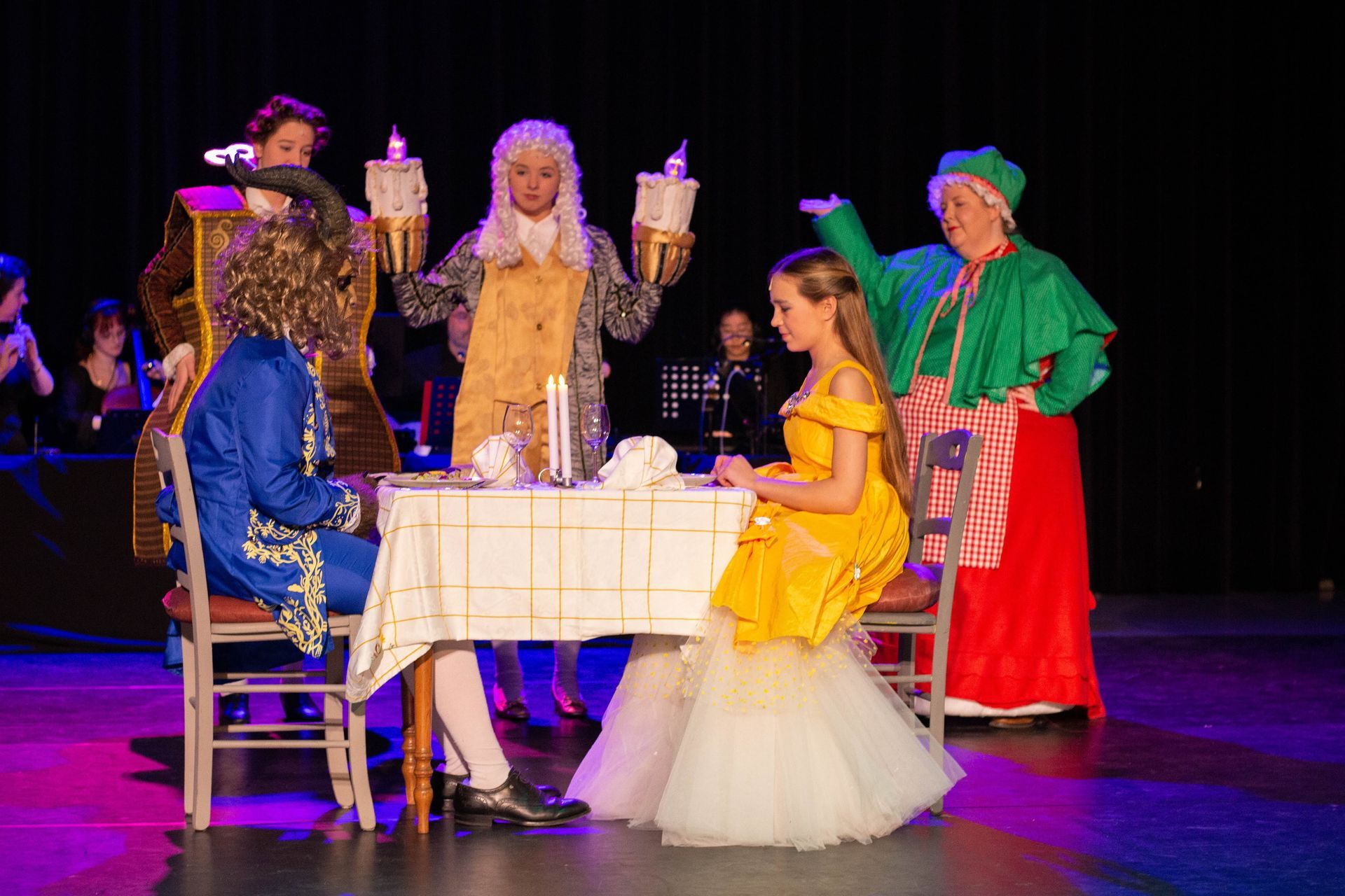 Toneelproductie van Belle en het Beest: Belle, het Beest, Lumière, Cogsworth en Mrs. Potts aan tafel.