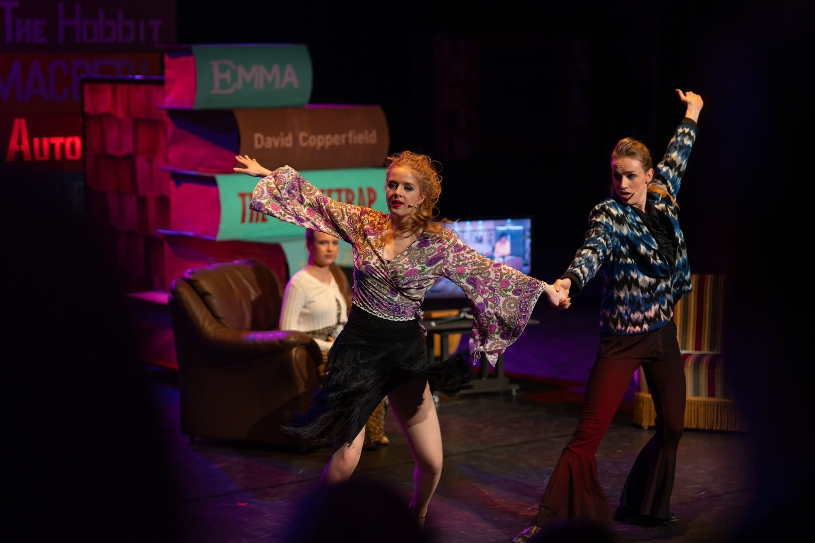 Twee dansers in kleding uit de jaren 70 staan ​​hand in hand op het podium en maken sprongen. Op de achtergrond: boeken, tv, een man in een stoel.
