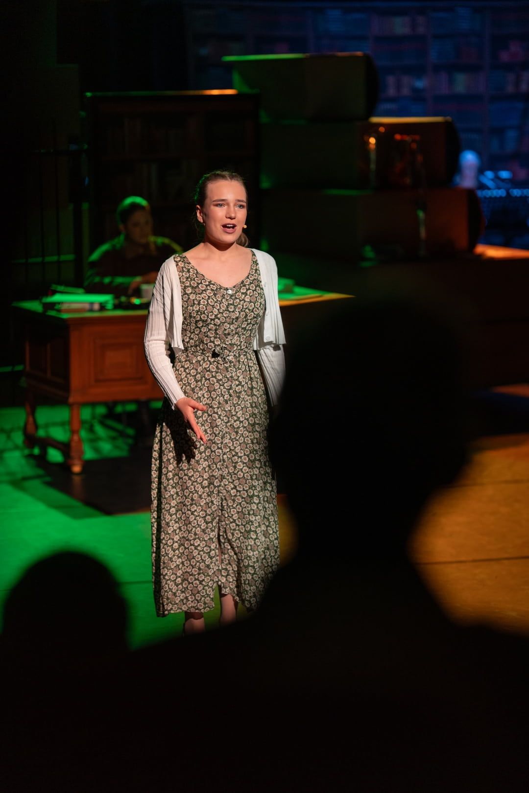 Een vrouw in een jurk zingt op het podium, met op de achtergrond iemand achter een bureau. Groen en geel licht.