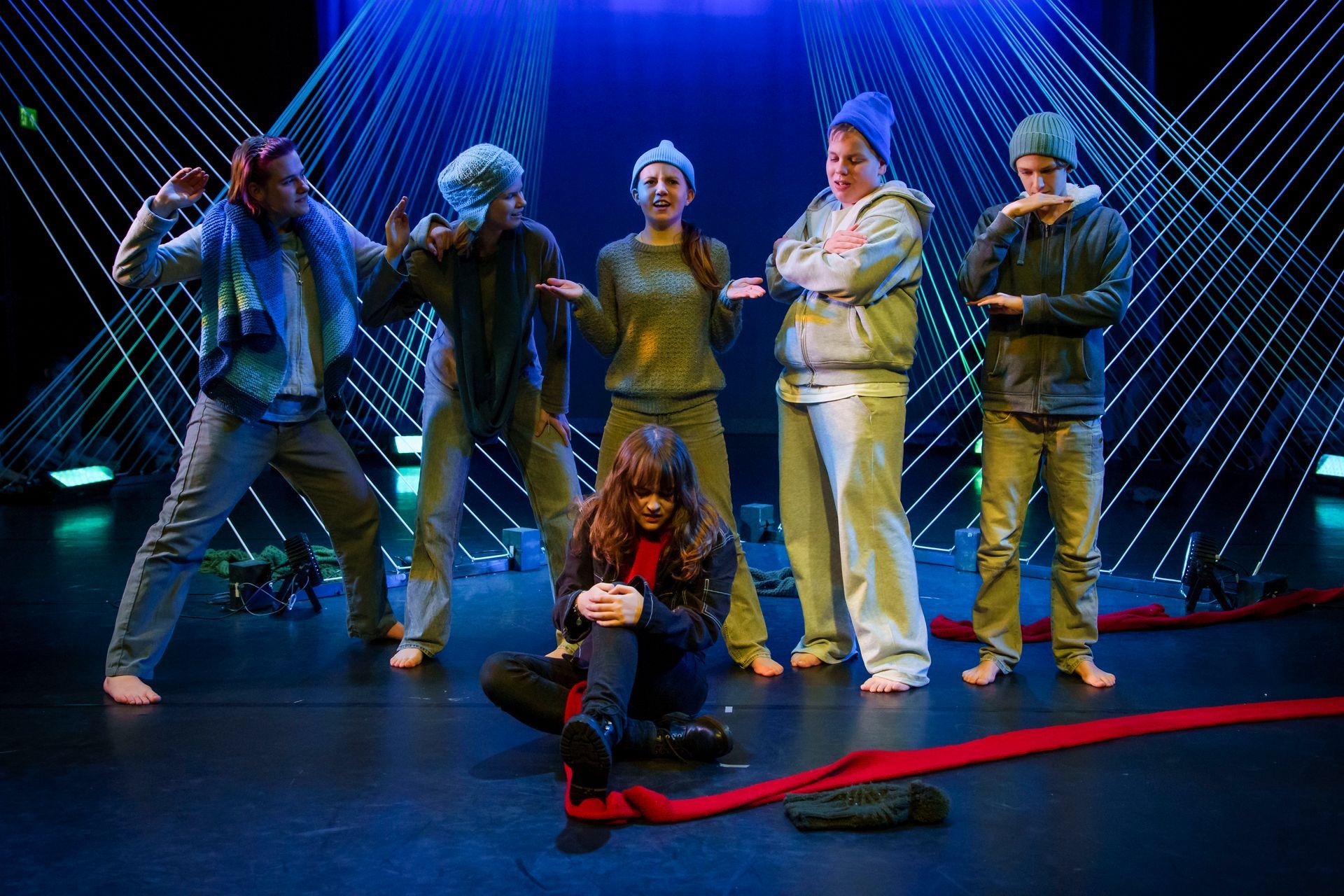 Scène uit een theaterproductie: Zes acteurs op het podium, één persoon zit en de anderen staan. Blauwe verlichting, rode sjaal.