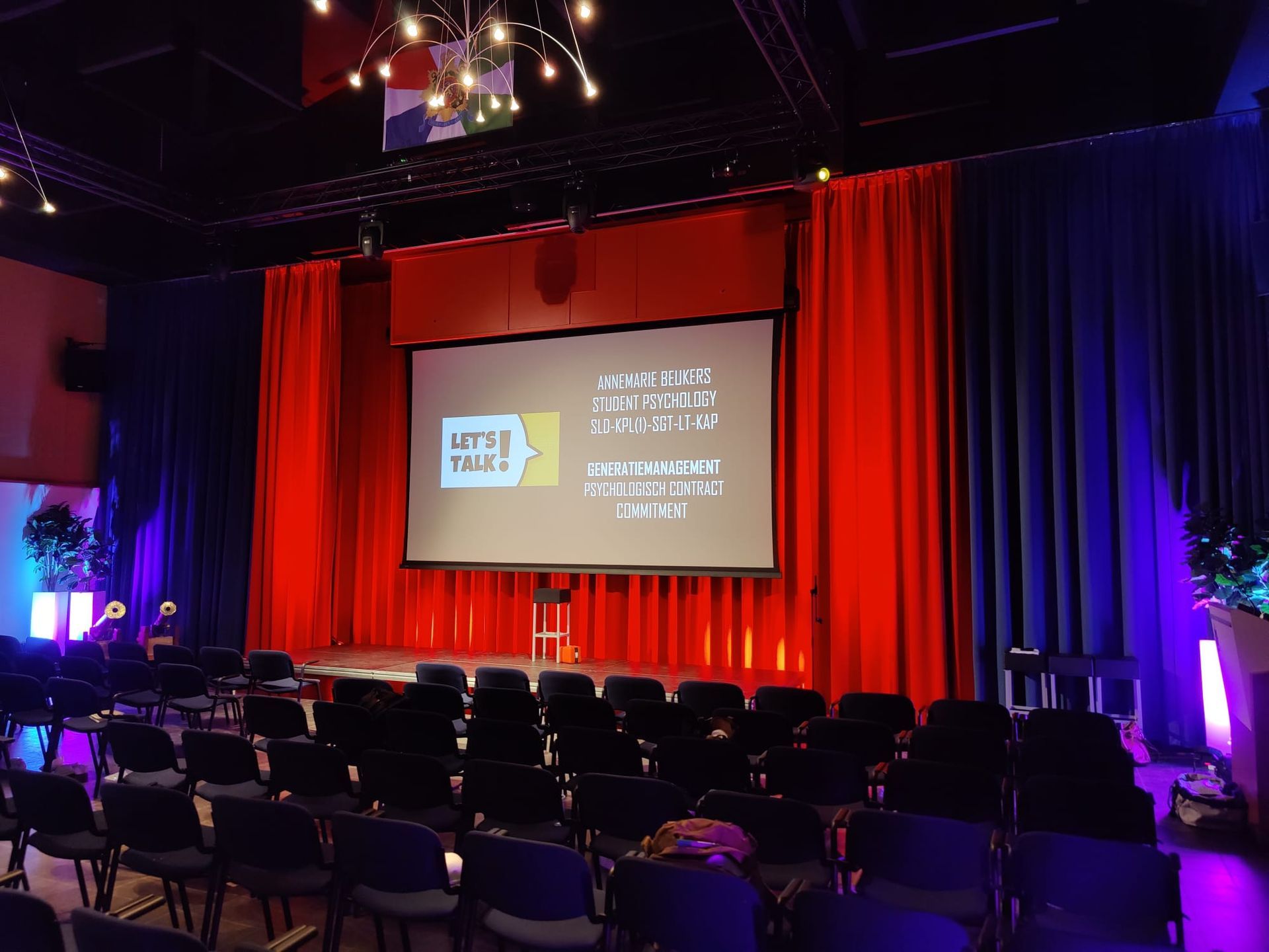 Een podium met een scherm, rode en blauwe gordijnen en rijen stoelen. Er is een presentatie aan de gang.