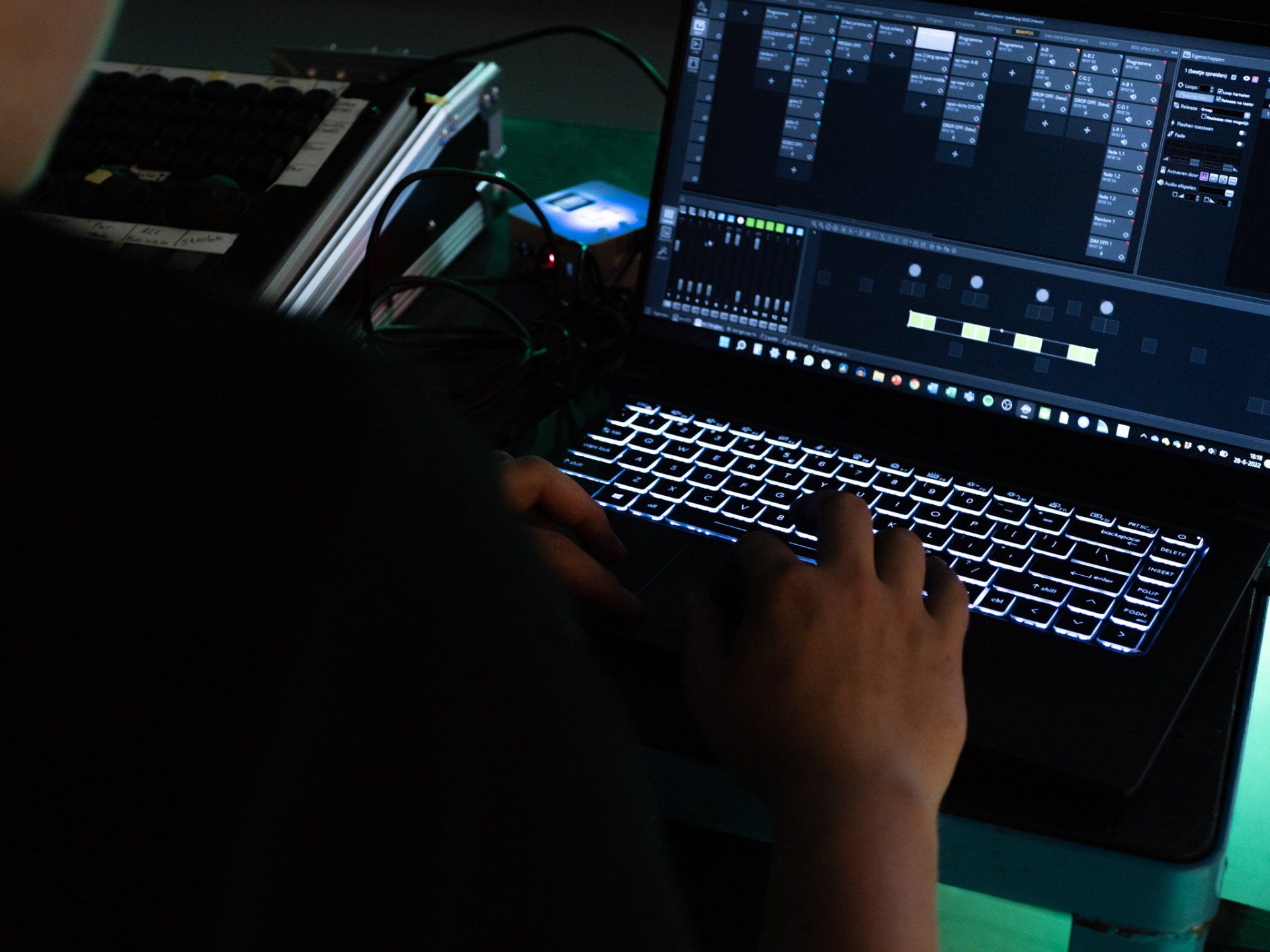 Persoon die een laptop met verlicht toetsenbord gebruikt om licht te bewerken; donkere omgeving met andere technische apparatuur.