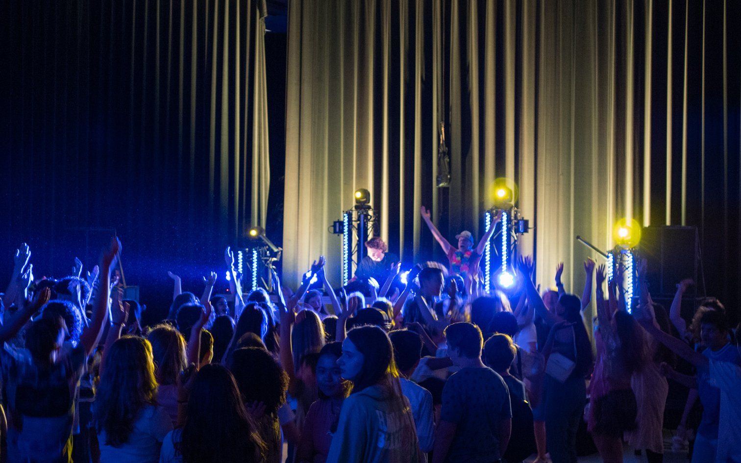 Concertpubliek met opgeheven armen, badend in blauw en geel podiumlicht. Een band treedt op het podium op.