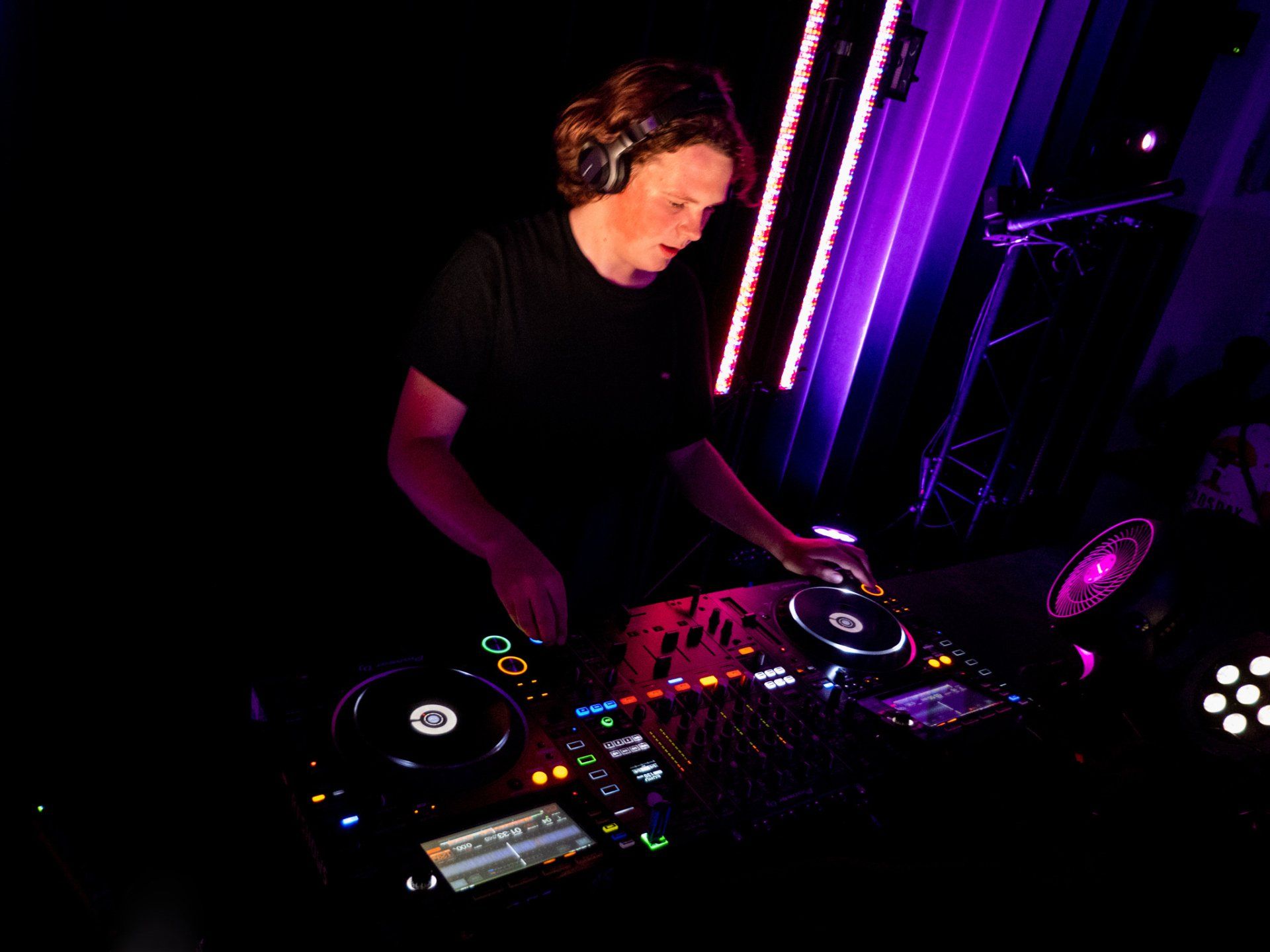 Een dj draait in een club, draagt ​​een koptelefoon, een donker shirt en gebruikt een mengpaneel, verlicht door paarse lampen.