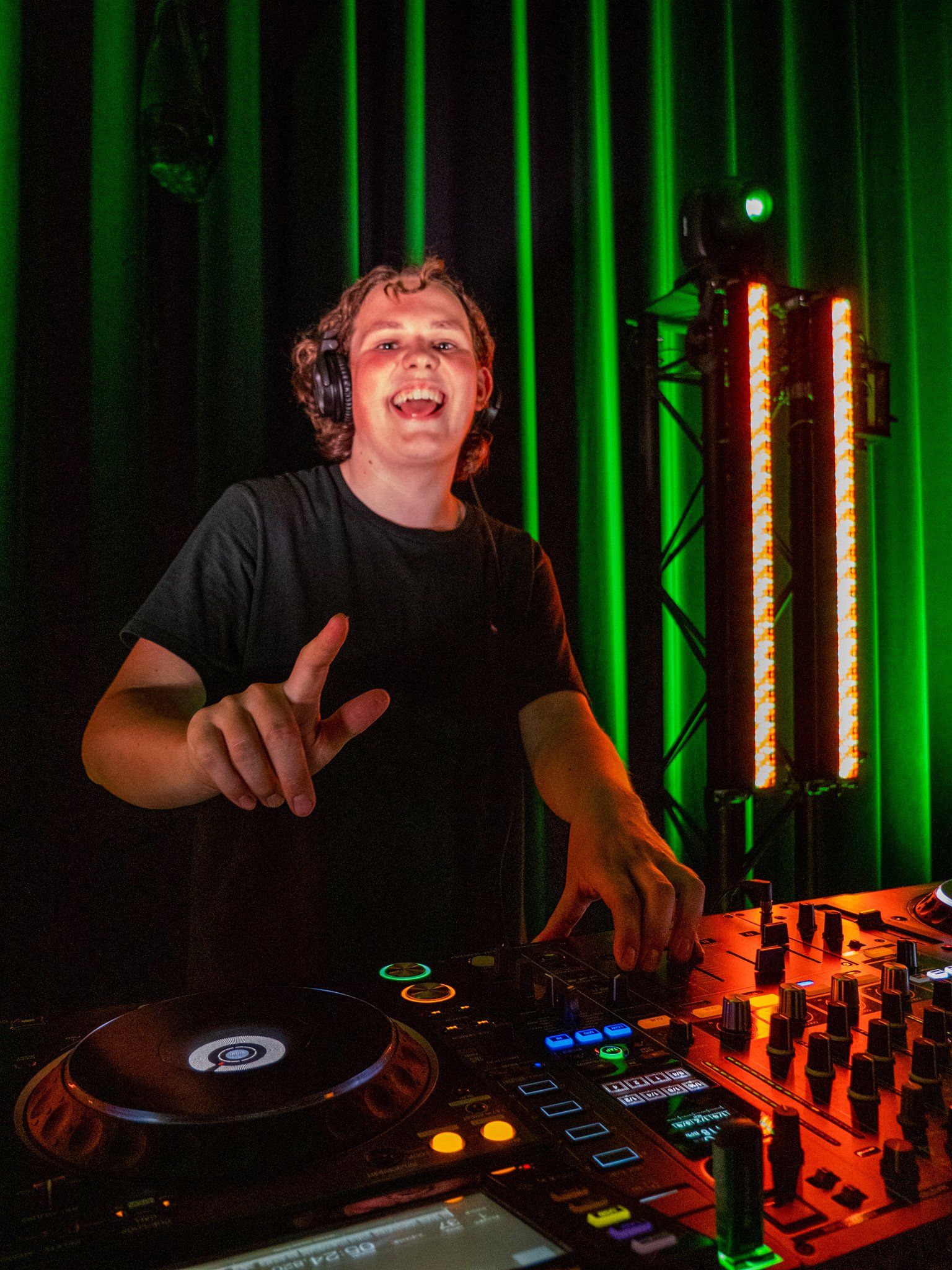 DJ in een muziekzaal, lachend en gebarend terwijl hij nummers mixt. Donker podium met groene achtergrond en verlichting.