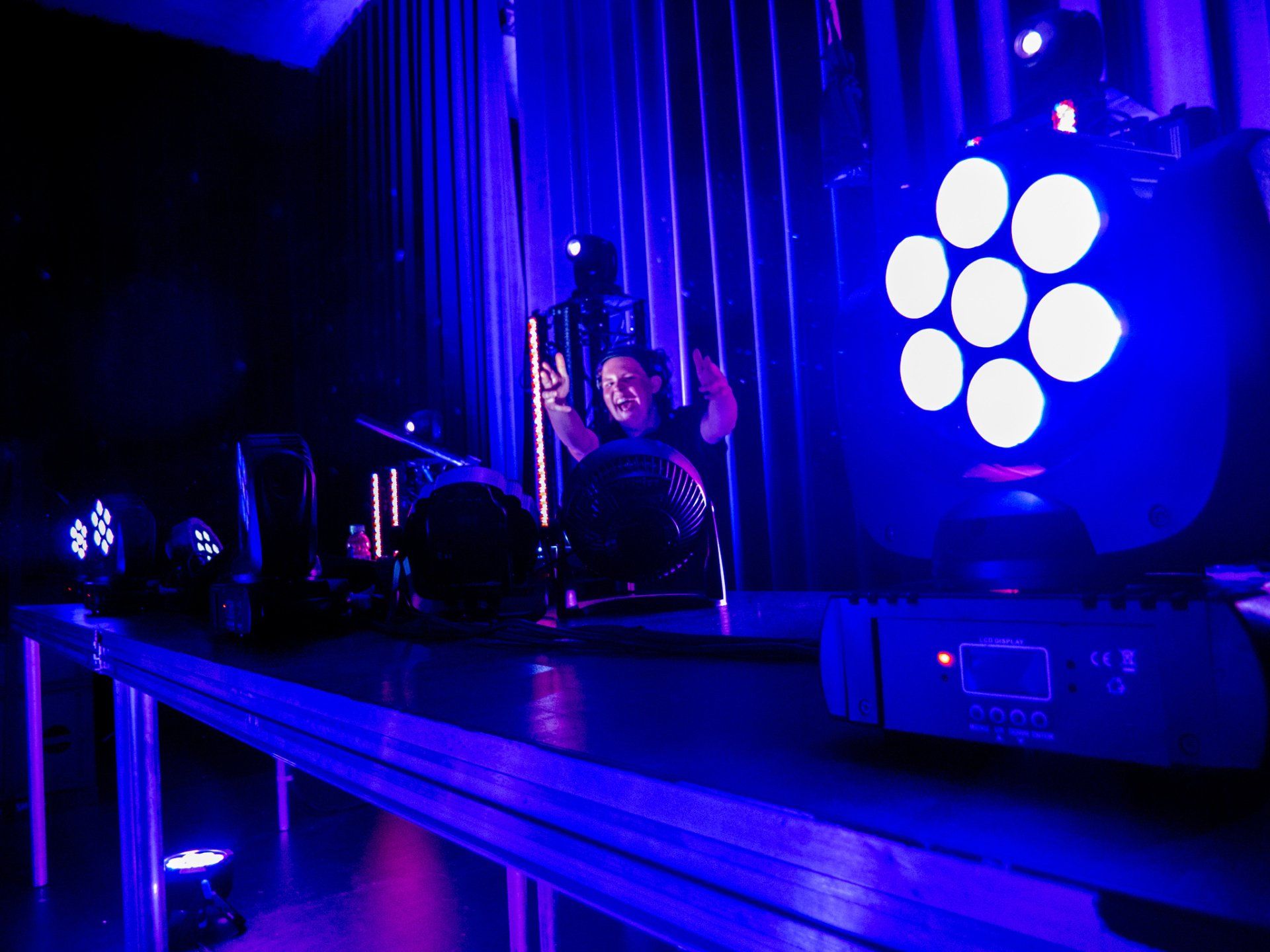 Een persoon, omringd door blauwe podiumverlichting, lachend, die een geluids- of lichttafel bedient.