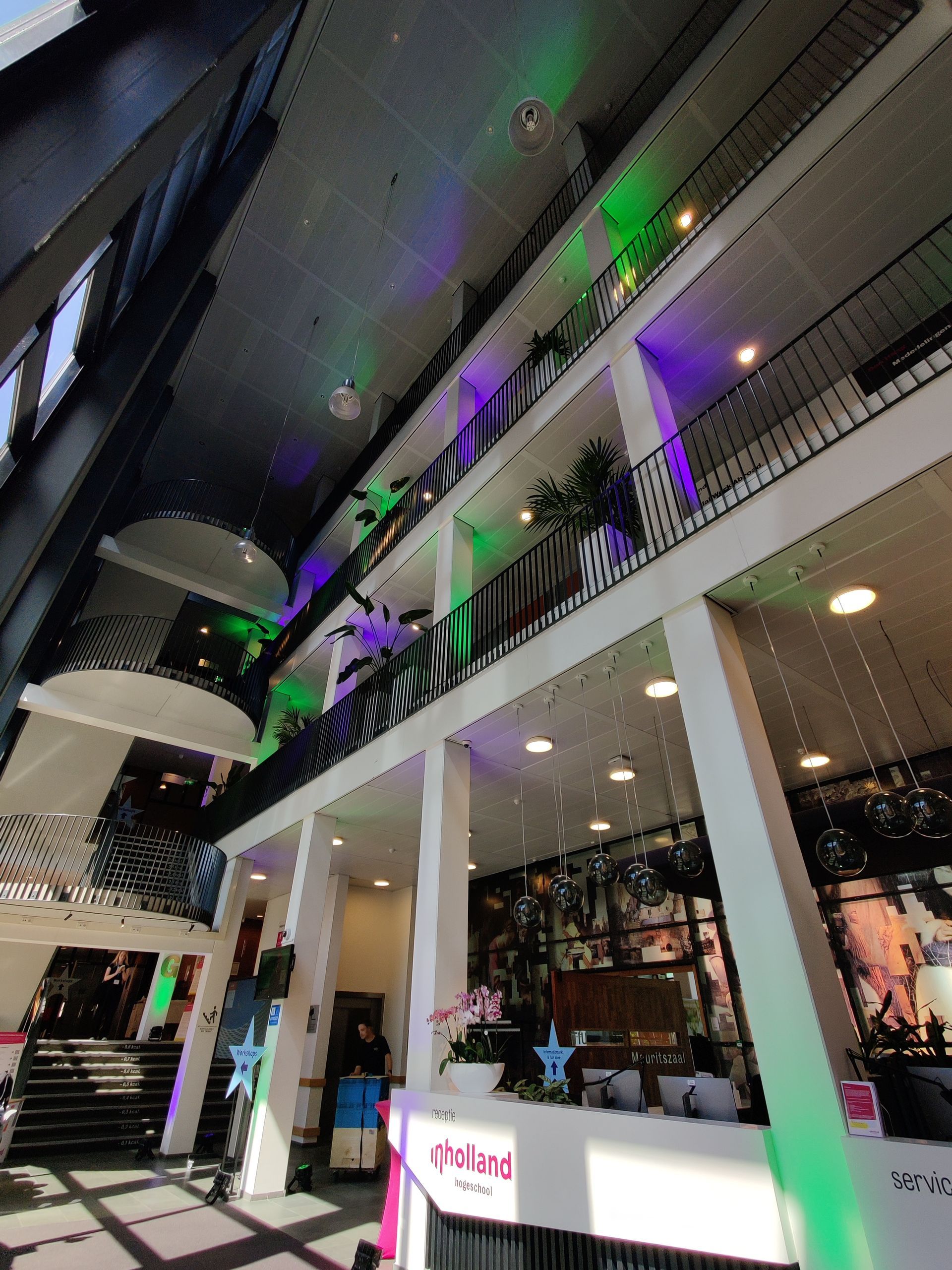 Interieur van een modern gebouw met meerdere verdiepingen, verlicht met groene en paarse lampen. De receptie is zichtbaar.