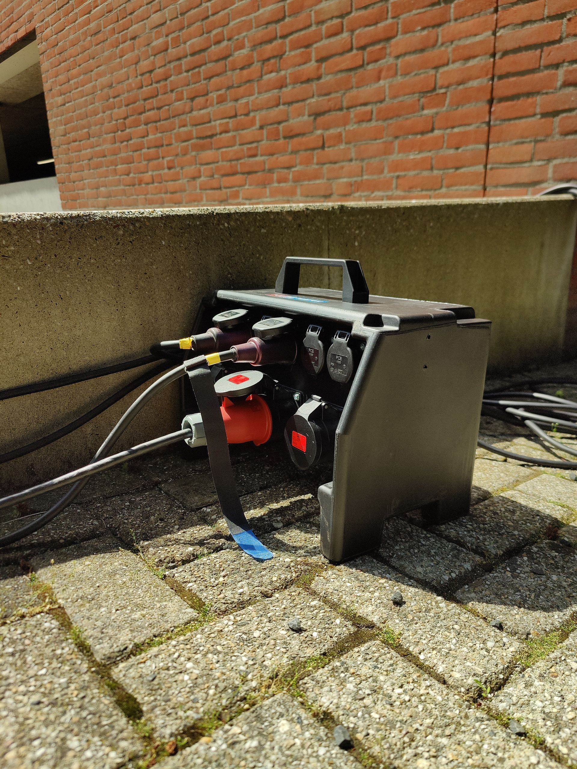 Zwarte stroomverdeelkast met diverse connectoren en kabels op bakstenen tegels naast een bakstenen muur.