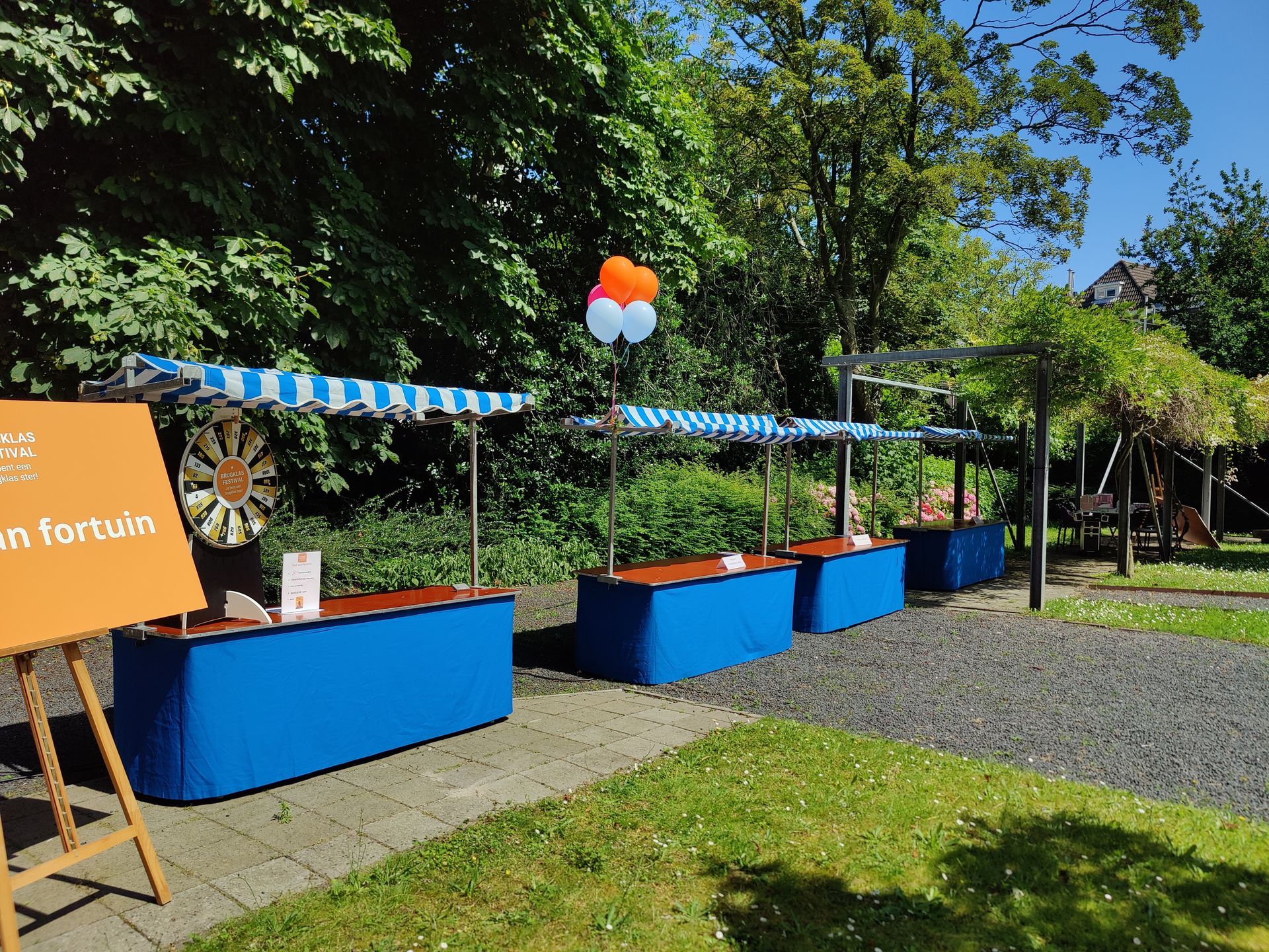 Drie blauwe verkoopkraampjes met gestreepte luifels in een park, met ballonnen erboven.