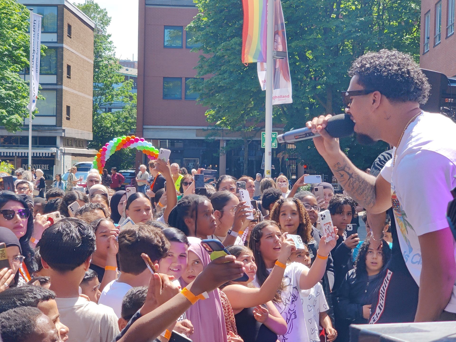 Zanger treedt op tijdens een openluchtevenement, een menigte kijkt toe, regenboogvlag op de achtergrond.