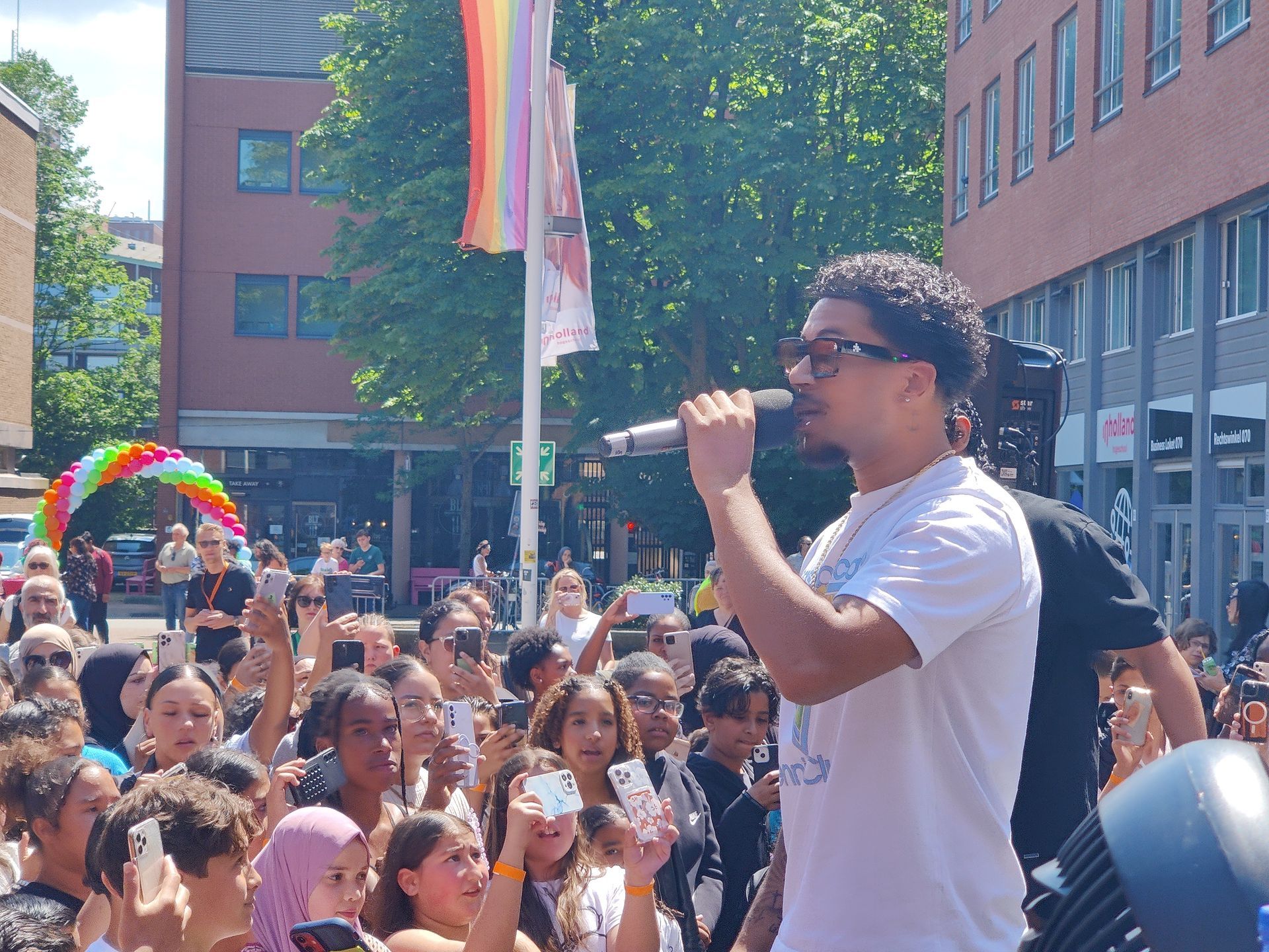 Een man zingt in een microfoon tijdens een openluchtevenement voor een grote menigte. Op de achtergrond is een regenboogvlag te zien.