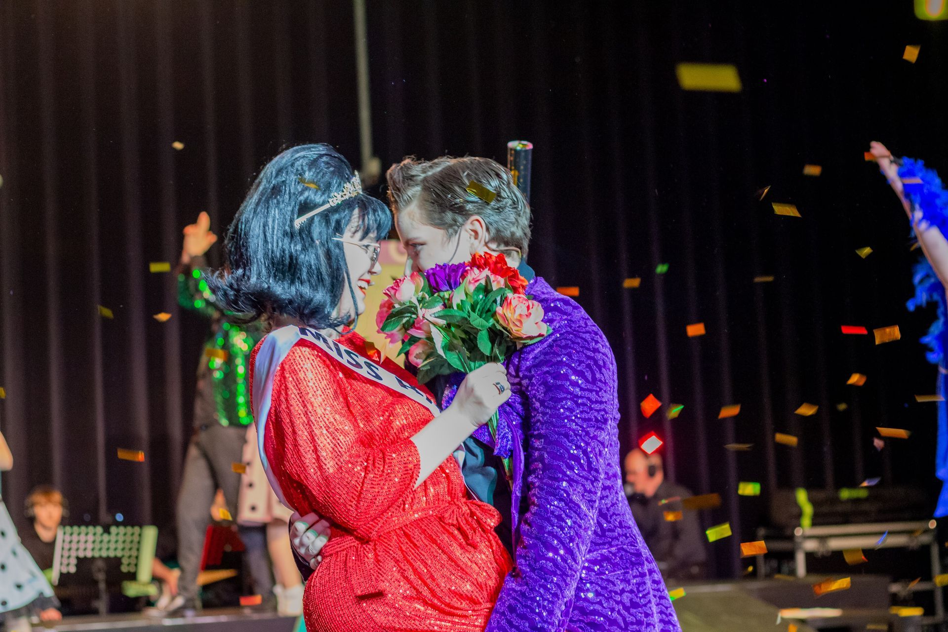 Een stel dat elkaar kust op het podium, rode jurk, paarse jas, confetti, boeket.