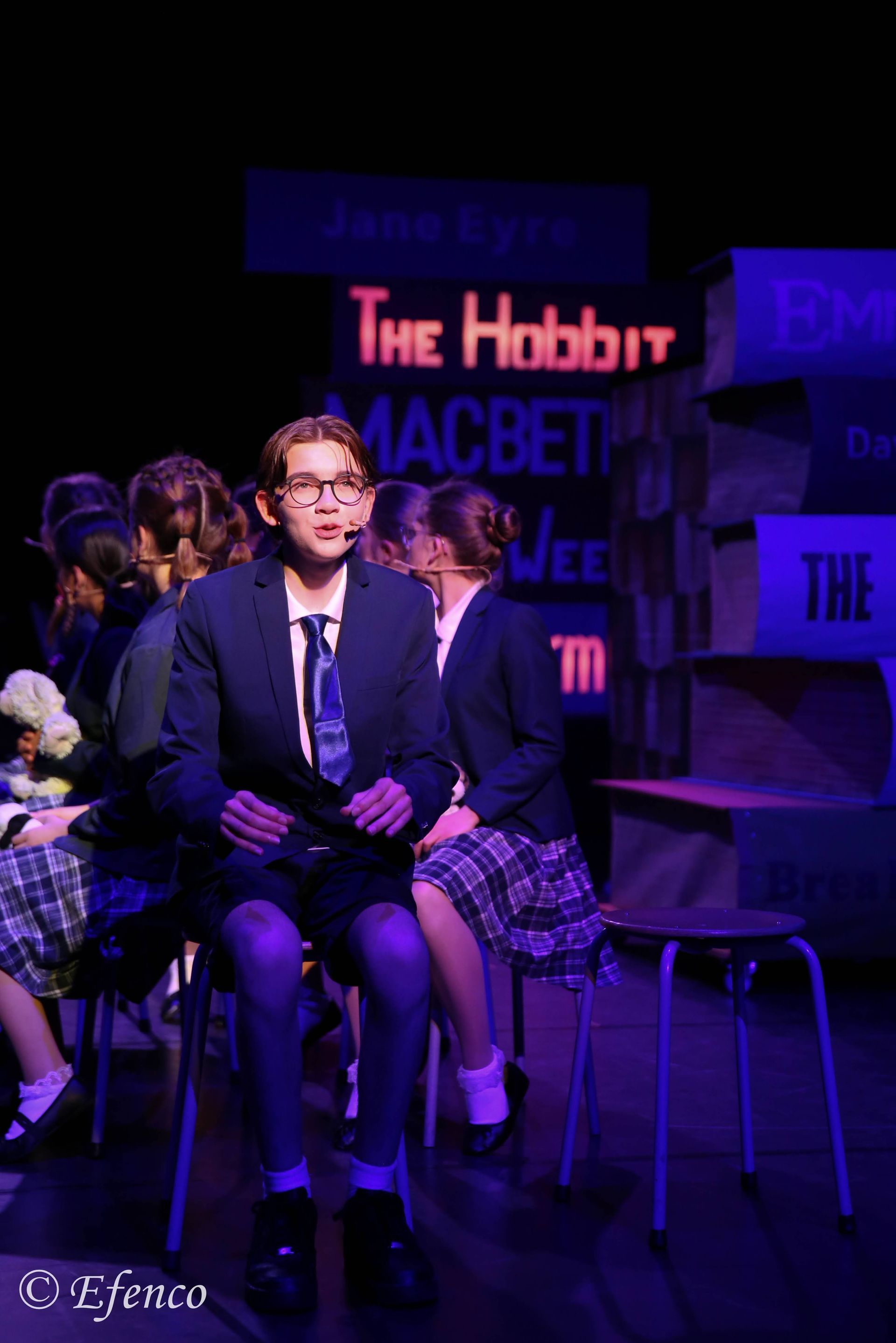 Een persoon in schooluniform zit op een stoel en spreekt op het podium; op de achtergrond zijn boektitels te zien.