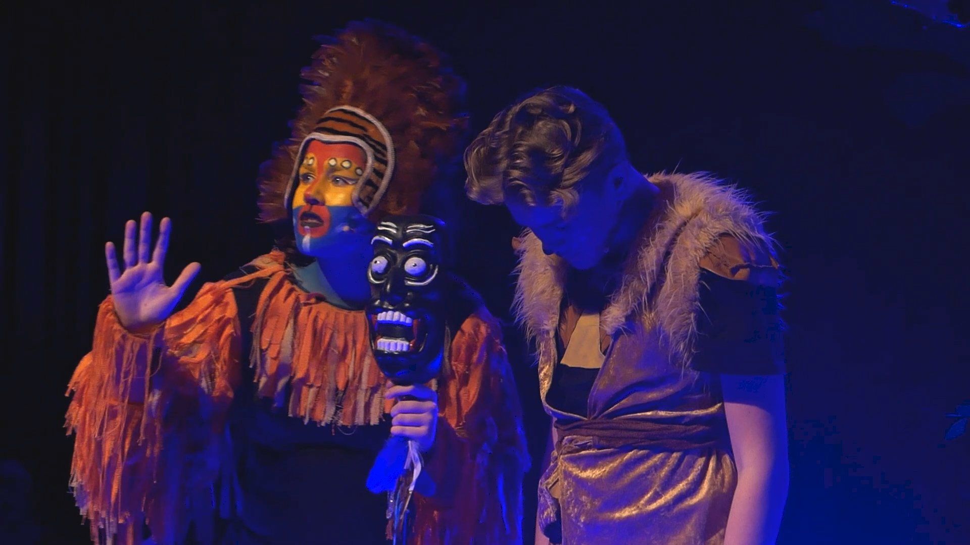 Acteurs in kostuums van de Leeuwenkoning op een podium. Een van hen houdt een stammenmasker vast en gebaart met zijn hand, de ander kijkt naar beneden.