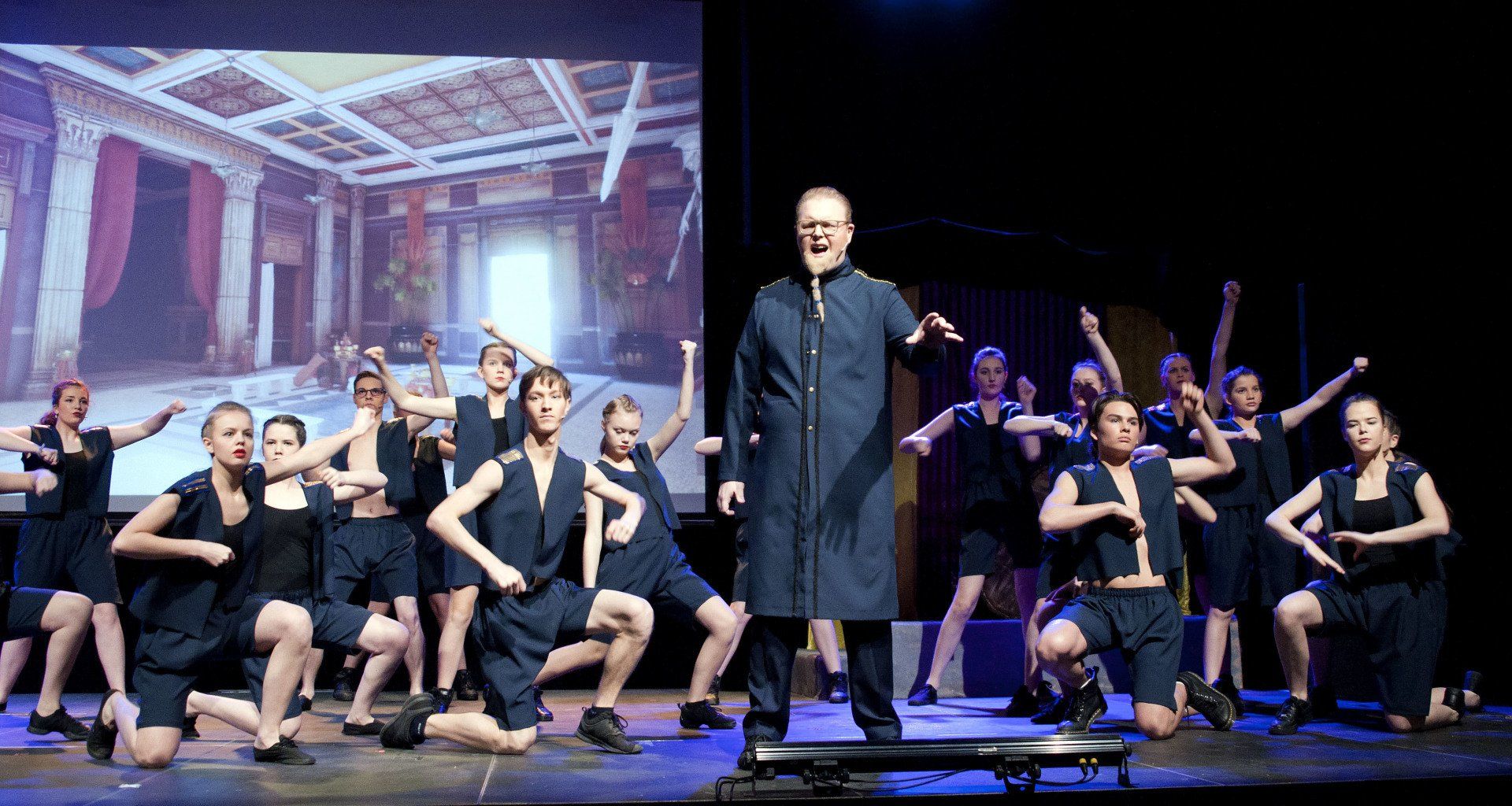 Een theatervoorstelling met een man in donkere gewaden en een groep dansers in donkere kostuums op het podium.