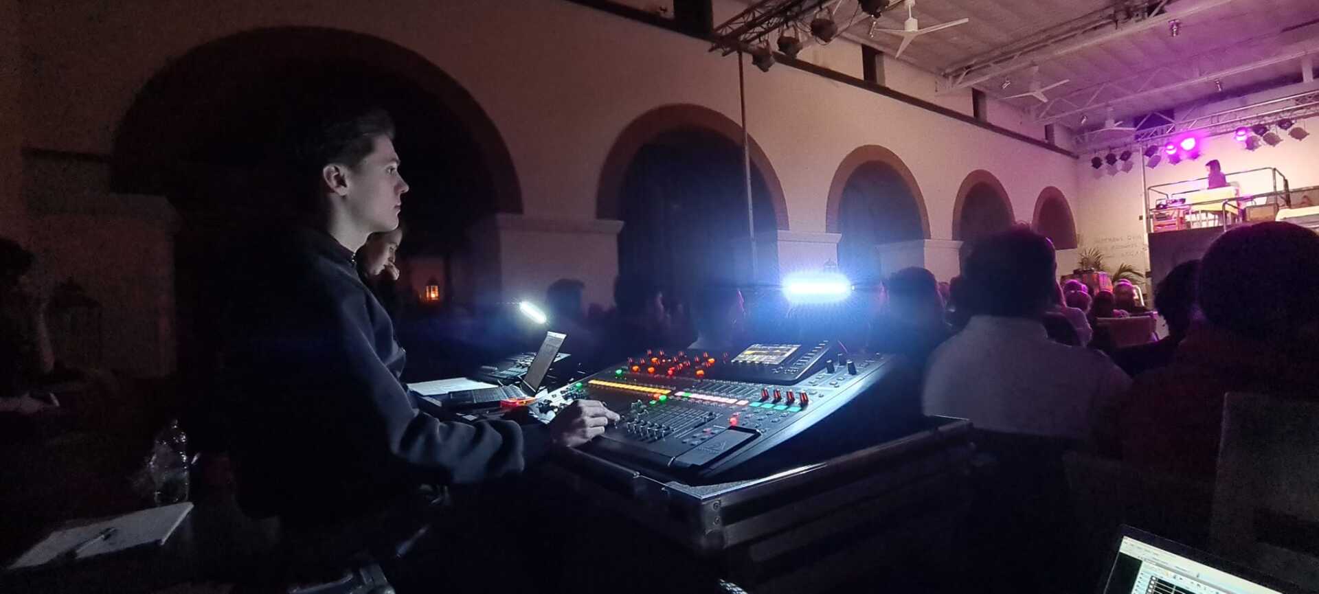 Iemand die de geluidsinstallatie bedient in een concertzaal, met het publiek op de achtergrond.