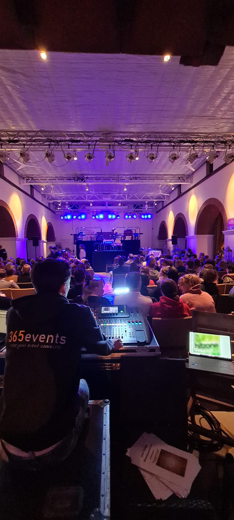 Een persoon staat achter een geluidsmixer in een evenementenhal met een publiek en een podium dat verlicht wordt met paarse lampen.